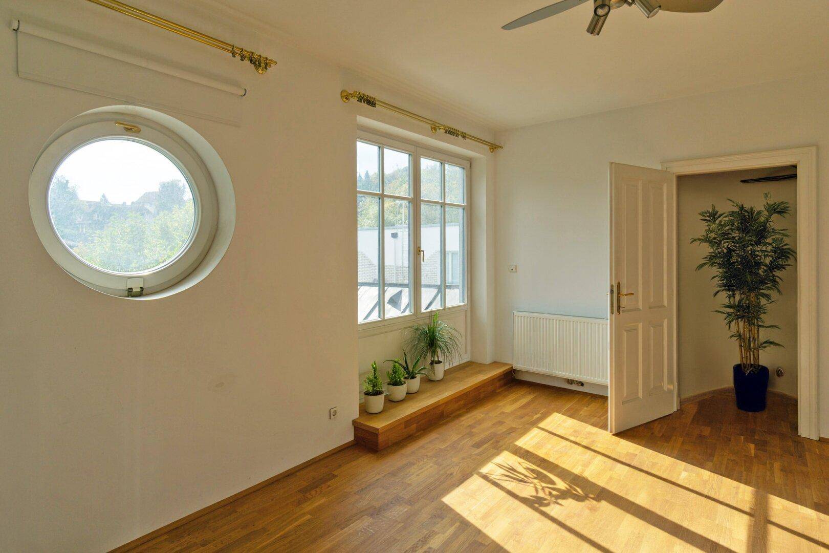 Helles Zimmer mit Holzboden, runden und rechteckigen Fenstern und viel Tageslicht.