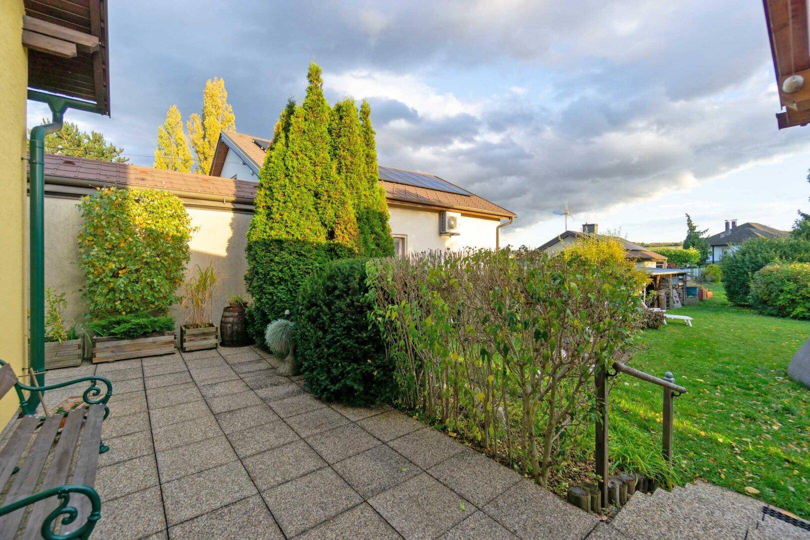 Gepflasterter Gartenbereich mit Sitzbank, Hecken und Blick auf die Umgebung.