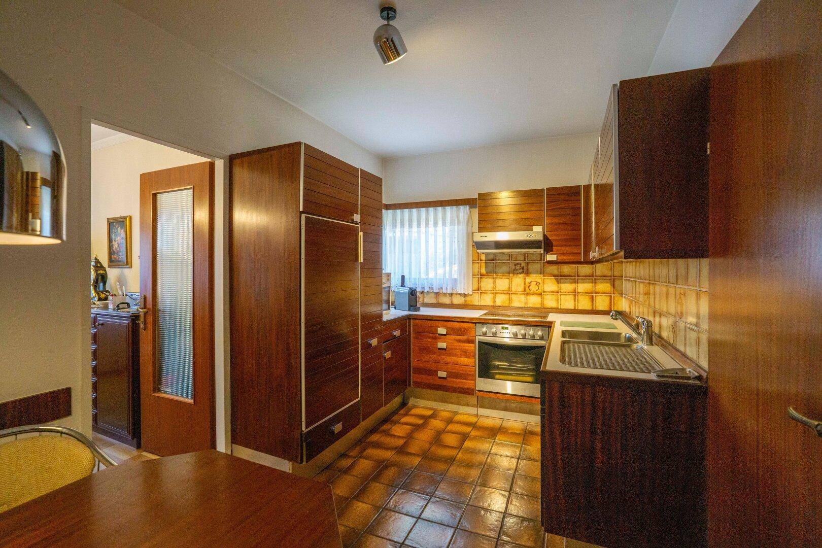 Traditionelle Küche mit Holzschränken, Fliesenboden und Fenster mit Blick ins Grüne.