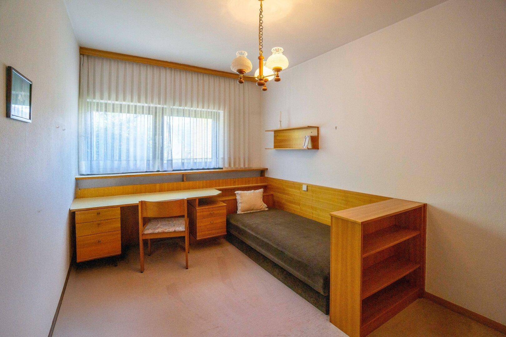 Helles Schlafzimmer mit Einzelbett, Schreibtisch und großem Fenster mit Vorhängen.