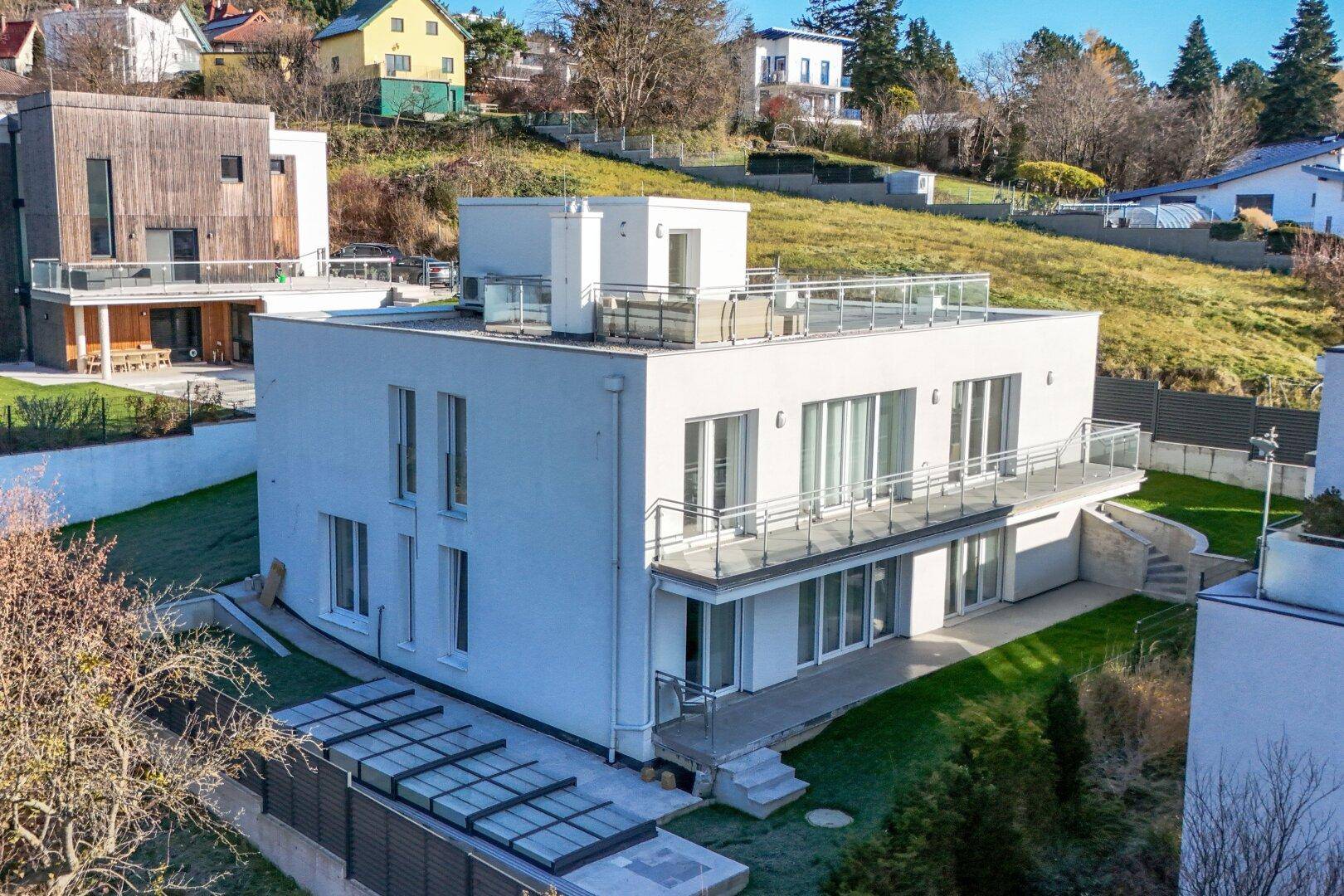 Modernes weißes Haus mit Flachdach, großen Fenstern und mehreren Balkonen in Hanglage.
