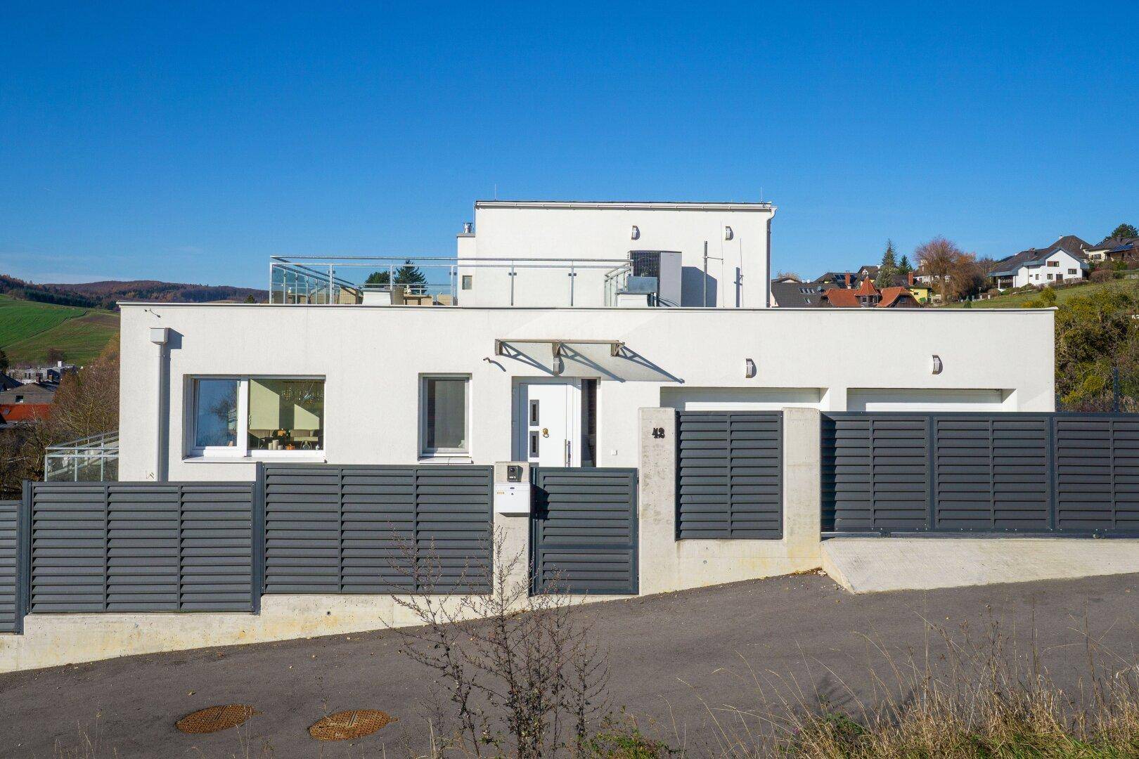 Moderne Hausfassade mit Eingangsbereich, Garagentoren und einem Zaun in Hanglage.