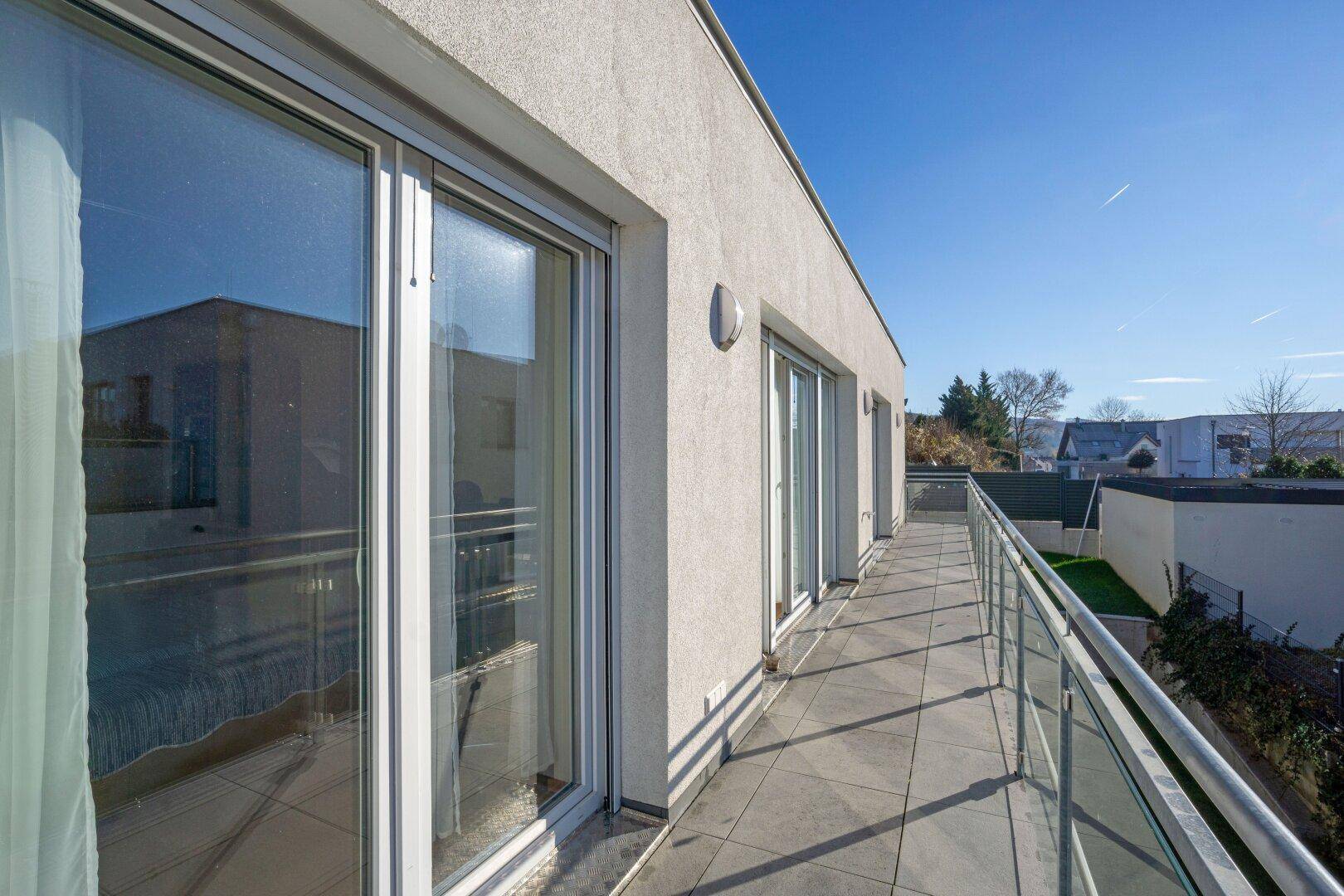 Langer Balkon mit Fliesenboden und Glasgeländer, zugänglich durch große Schiebetüren.