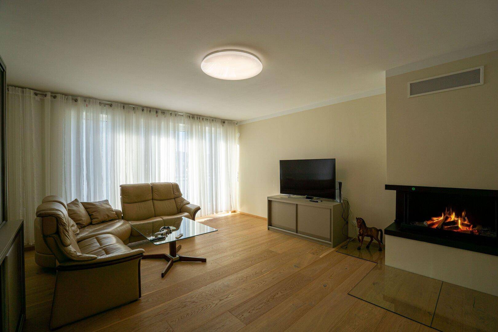 Gemütliches Wohnzimmer mit Parkettboden, Kamin und großen Fenstern mit Vorhängen.