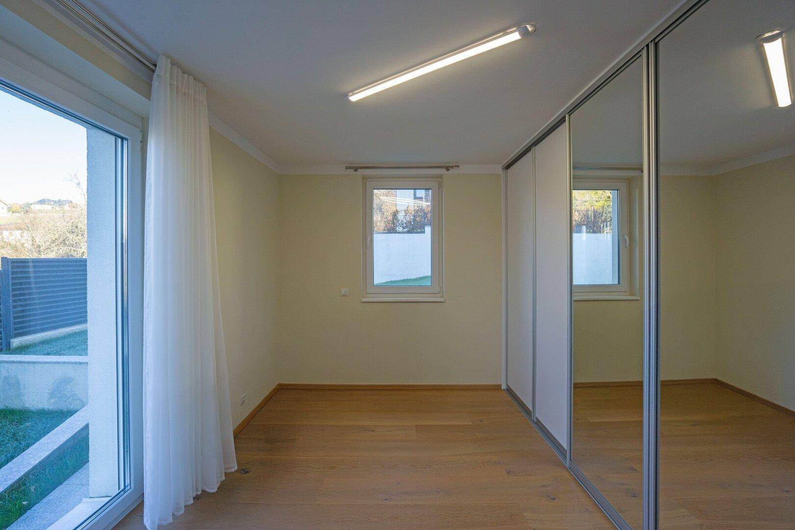 Leeres Zimmer mit Parkettboden, großen Fenstern und viel Tageslicht.