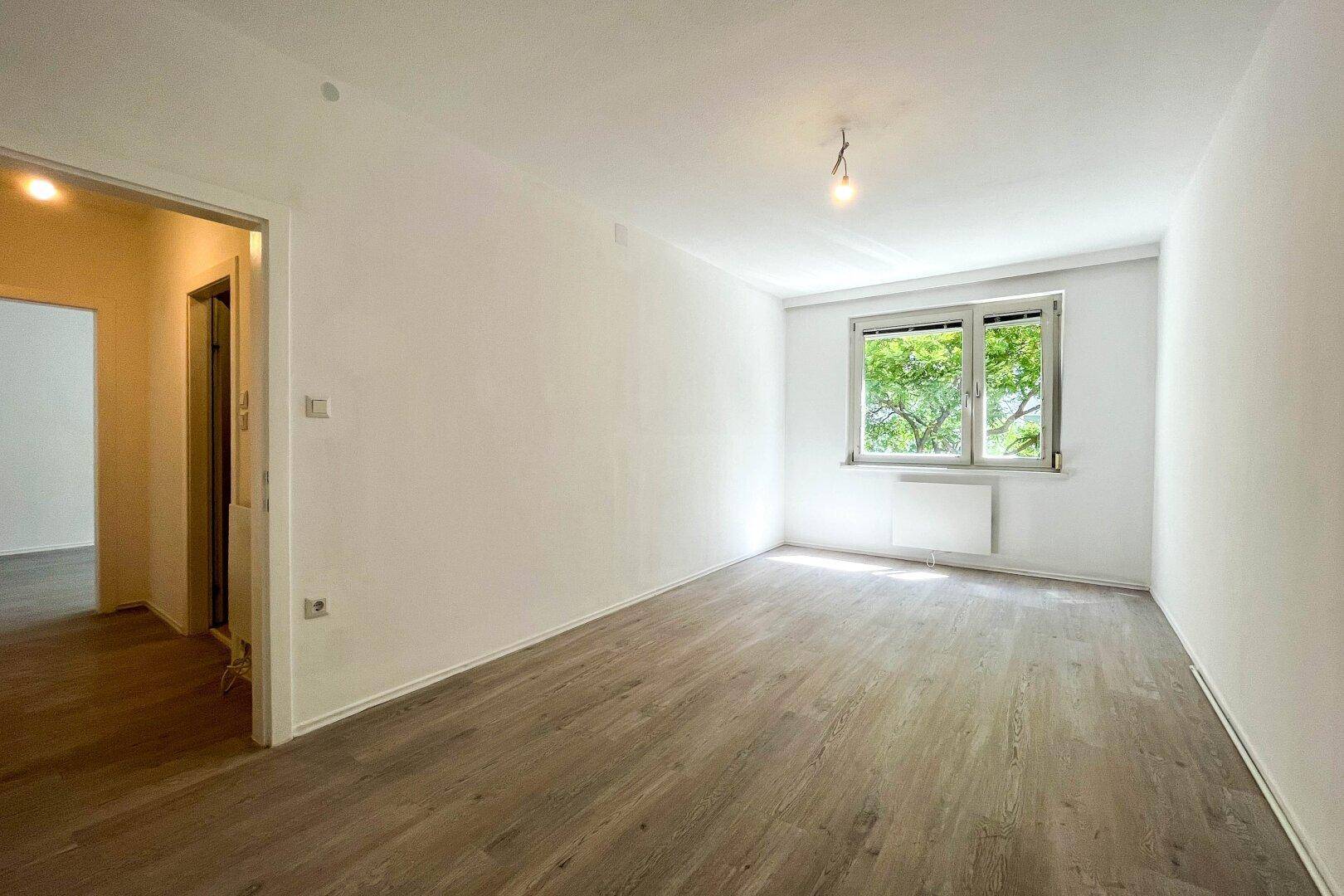 Geräumiges, helles Zimmer mit Laminatboden und Blick auf Bäume.
