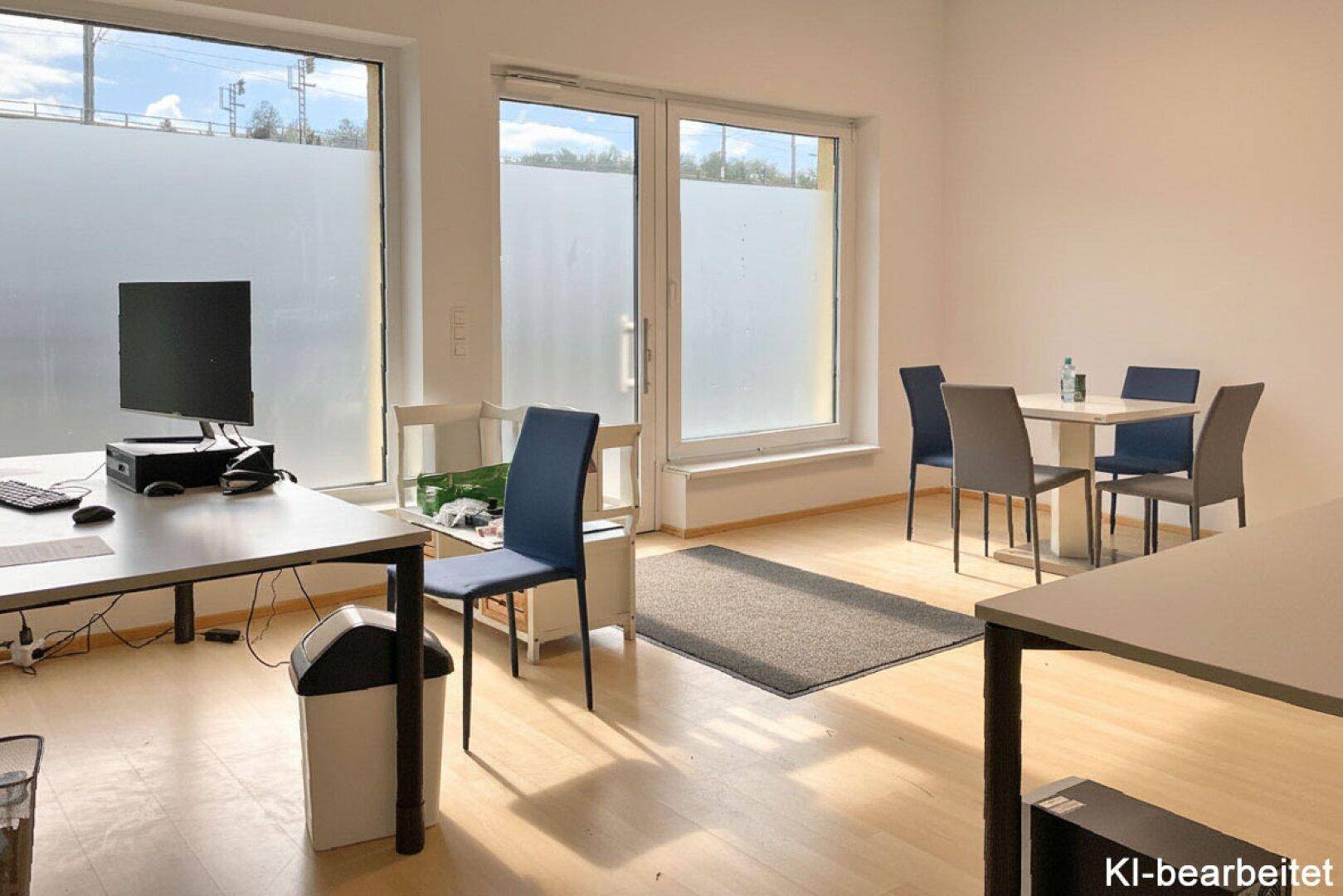 Heller Büroraum mit großen Fenstern, Arbeitsplätzen und einem kleinen Essbereich mit modernen Möbeln.