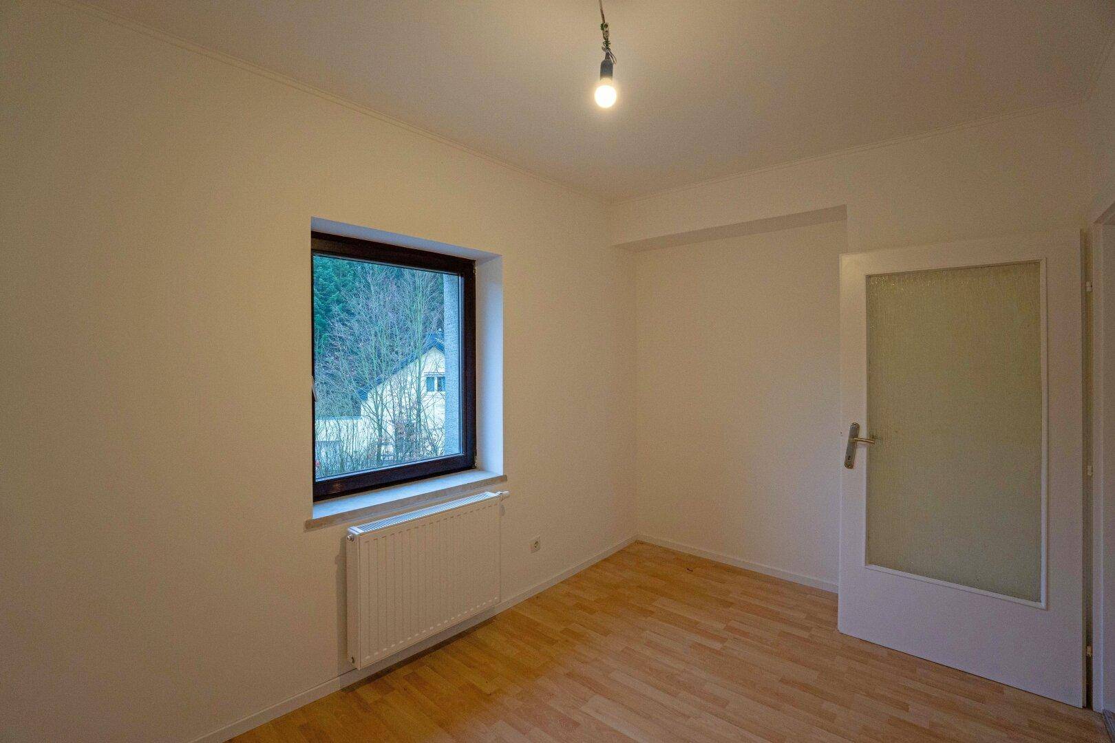 Helles Schlafzimmer mit Laminatboden und einem Fenster, das den Blick ins Grüne freigibt.