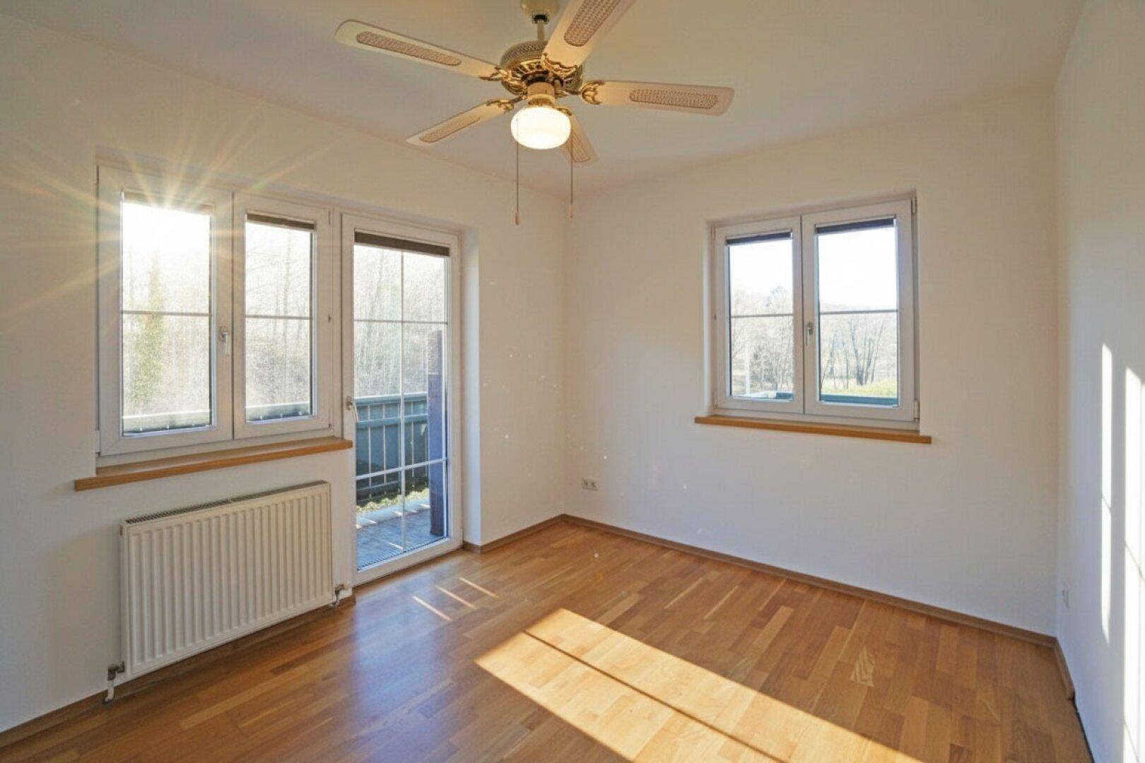 Sonniges Schlafzimmer mit Parkettboden, Deckenventilator und Fenstern mit Blick ins Grüne.