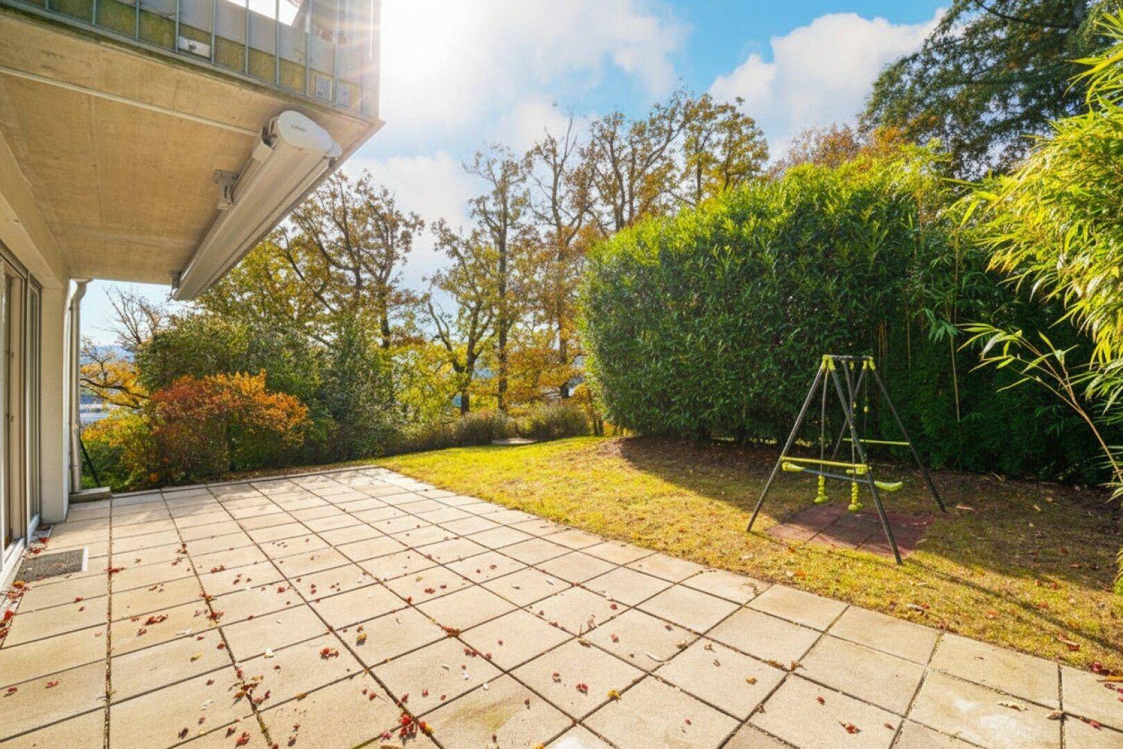Sonnige Terrasse mit Steinplatten, angrenzendem Gartenbereich und Kinderschaukel.