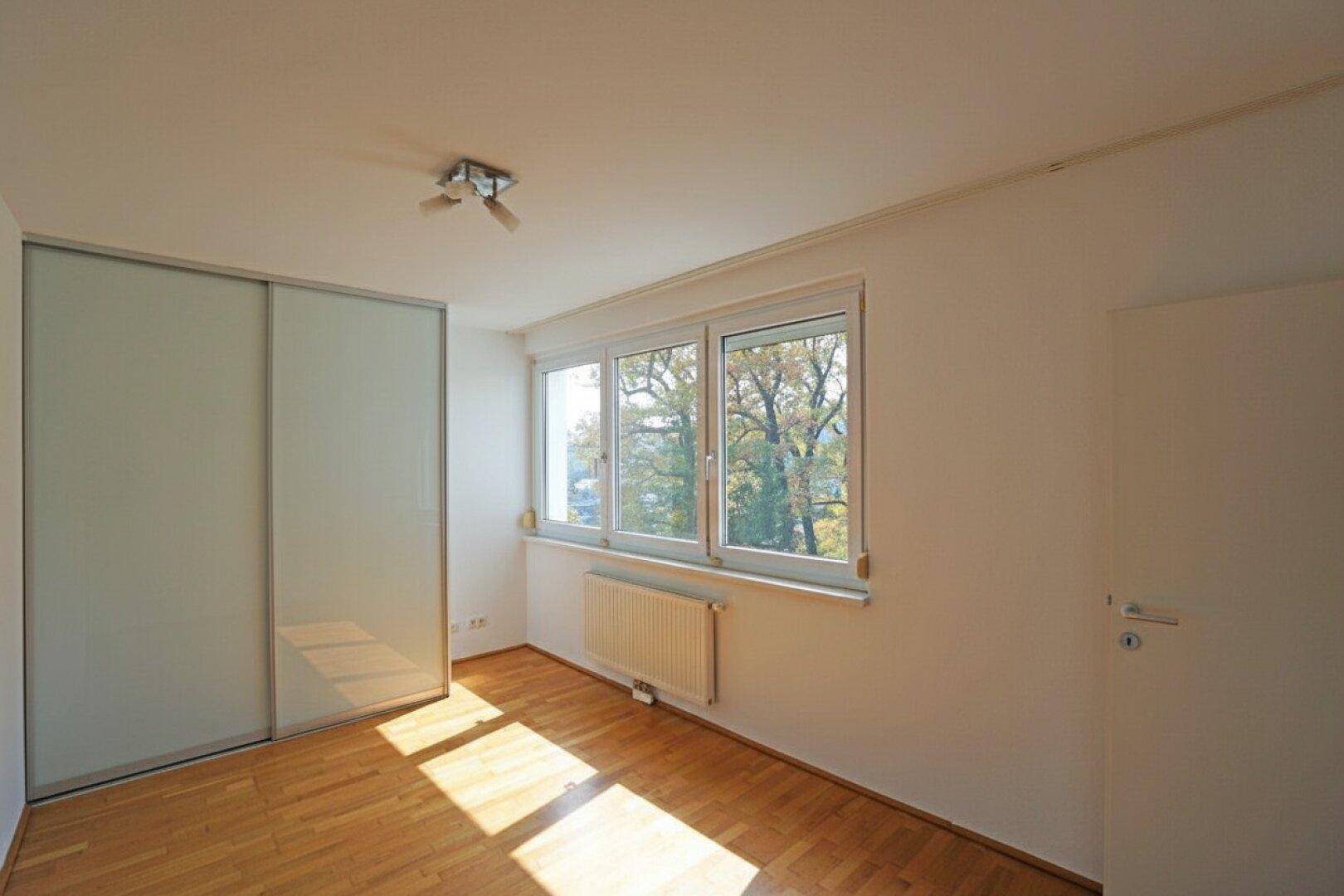 Helles Schlafzimmer mit Parkettboden, großem Fenster und praktischem Einbauschrank.