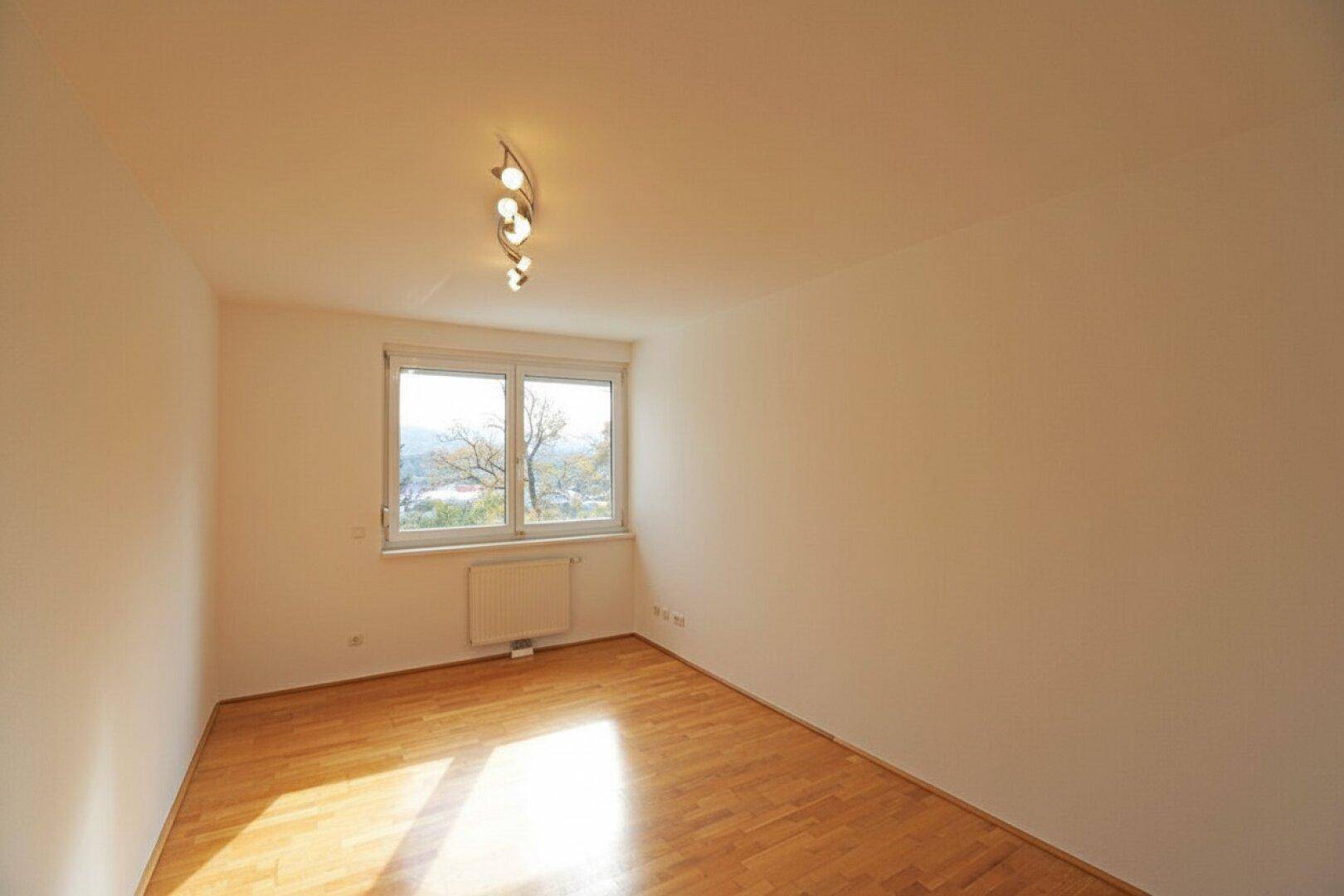 Lichtdurchflutetes Zimmer mit hellem Parkettboden und Blick ins Grüne durch das Fenster.