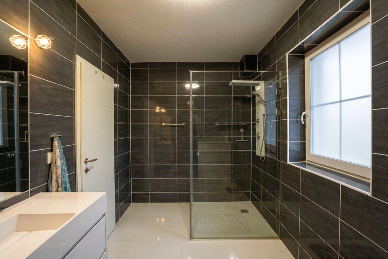 Modernes Badezimmer mit dunklen Wandfliesen, ebenerdiger Glasdusche und Fenster.