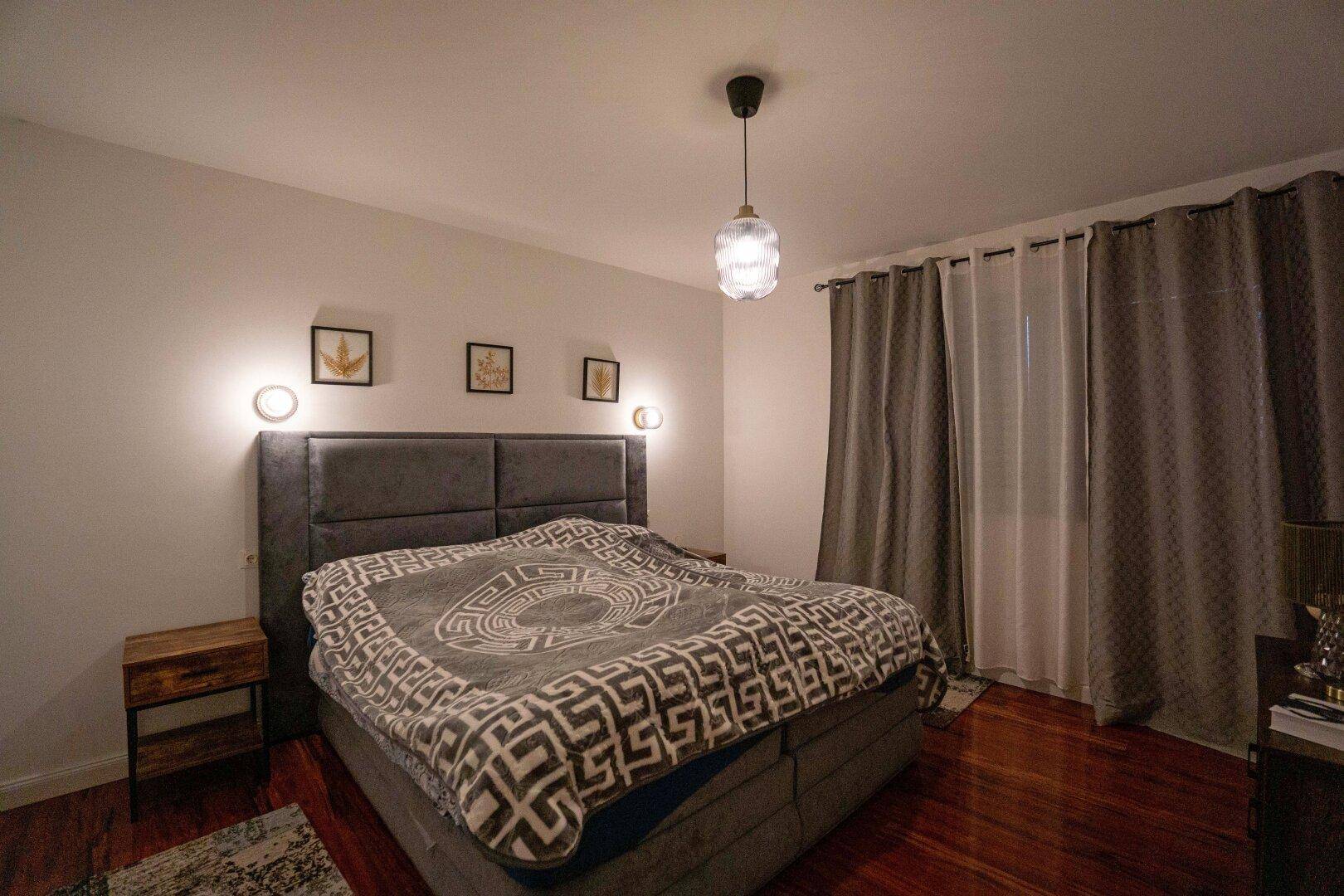 Gemütliches Schlafzimmer mit großem Polsterbett, dunklem Holzboden und Vorhängen.