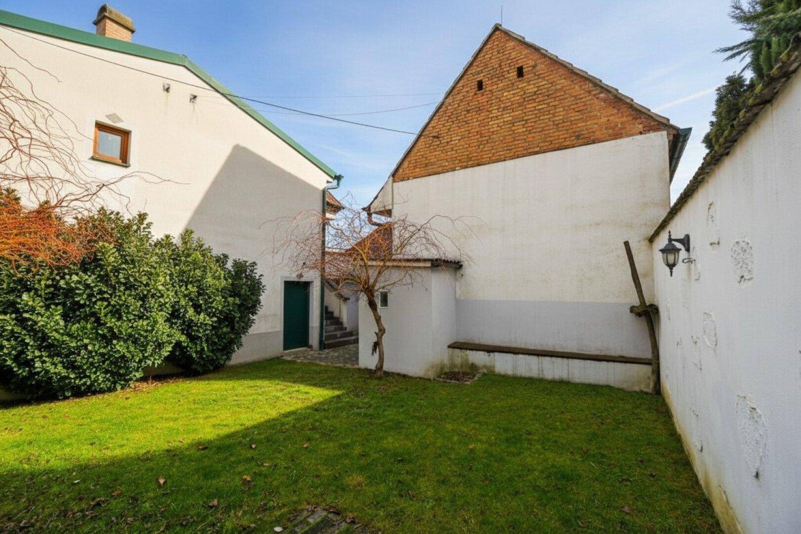 Hinterhofgarten mit Rasen, ummauertem Bereich und Zugang zu einem Nebengebäude.