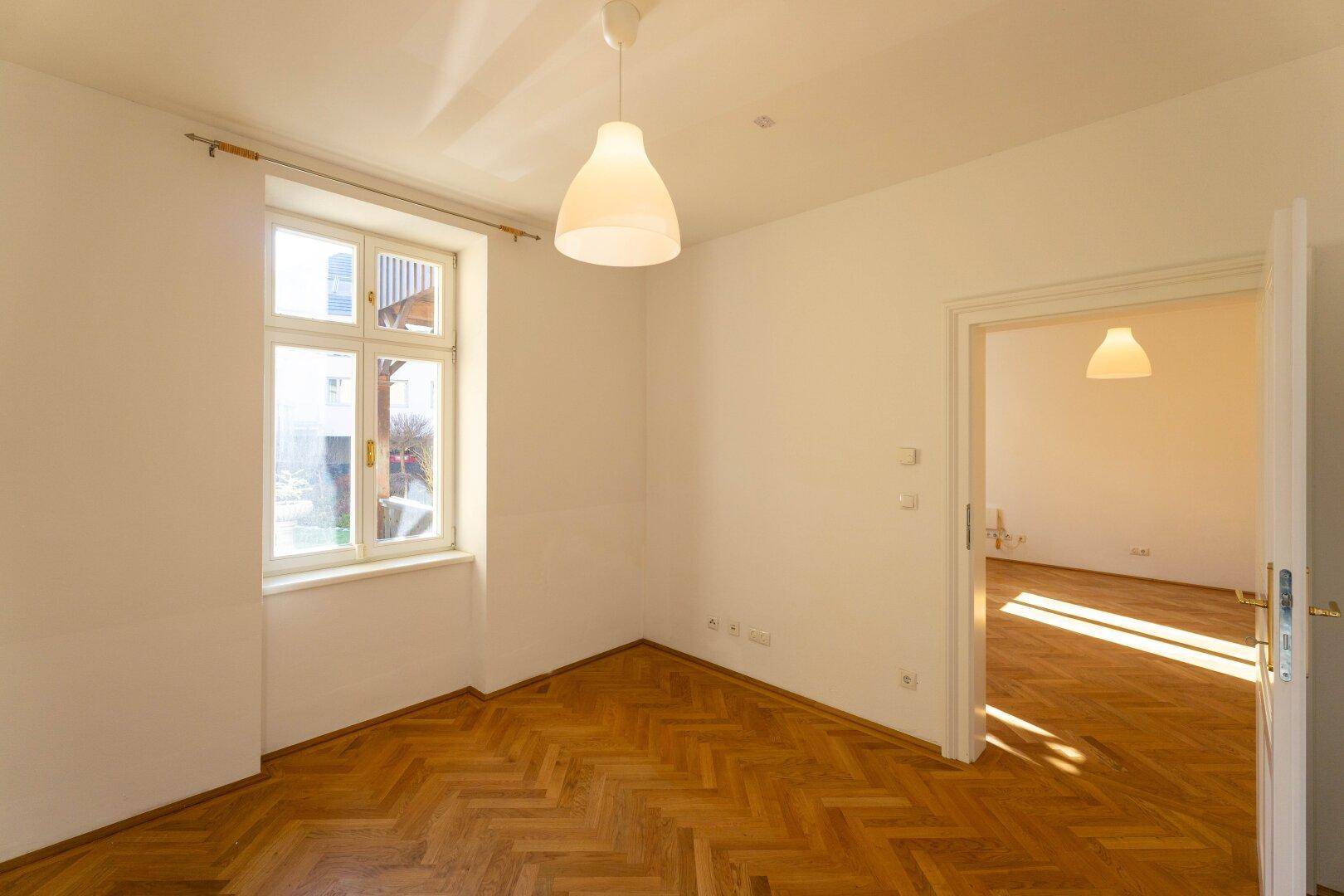 Leerer, sonniger Raum mit Fischgrätparkett und einem großen Fenster, das viel Tageslicht spendet.