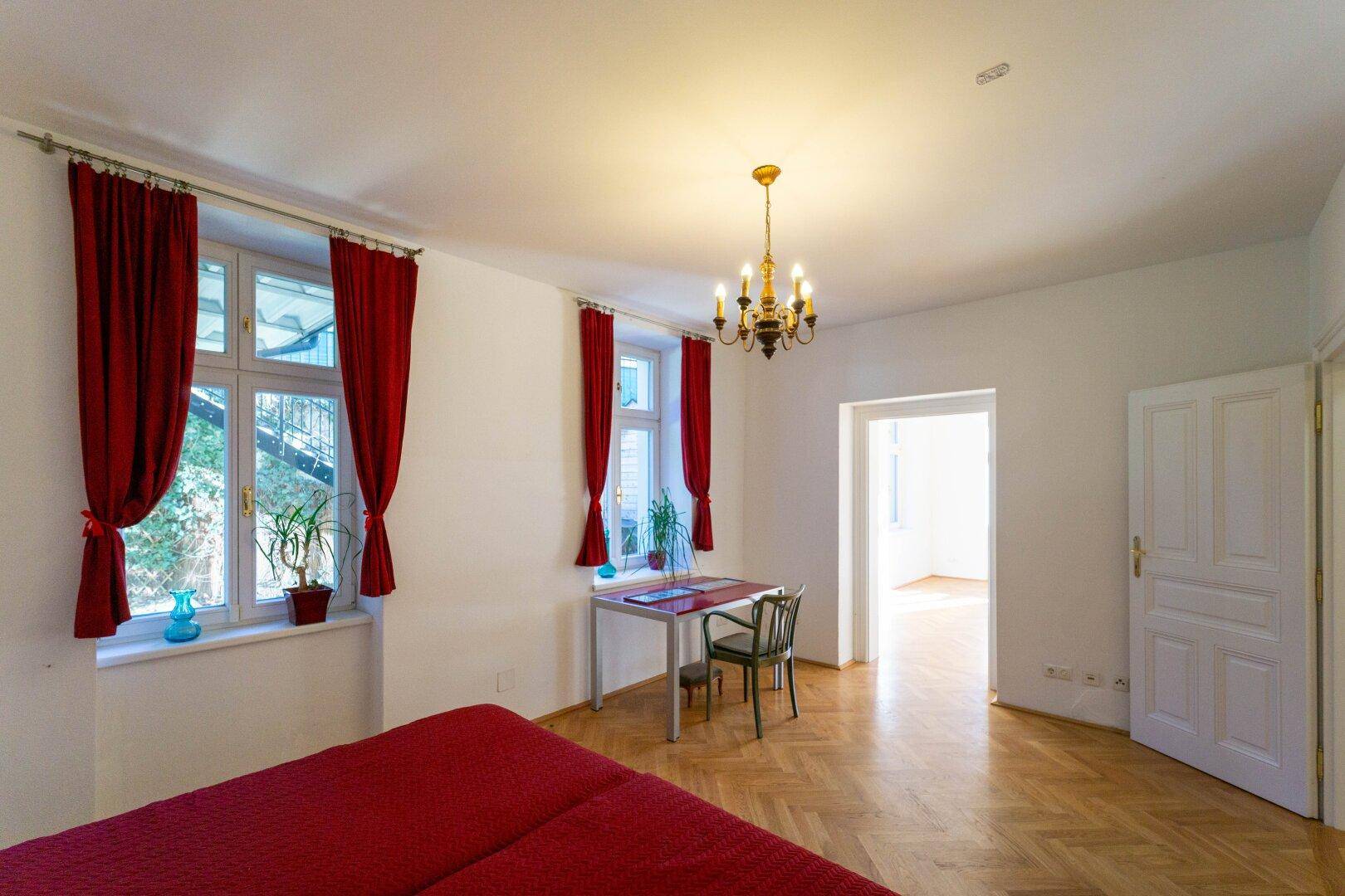 Helles Schlafzimmer mit Parkettboden, Bett und roten Vorhängen, das viel Tageslicht erhält.
