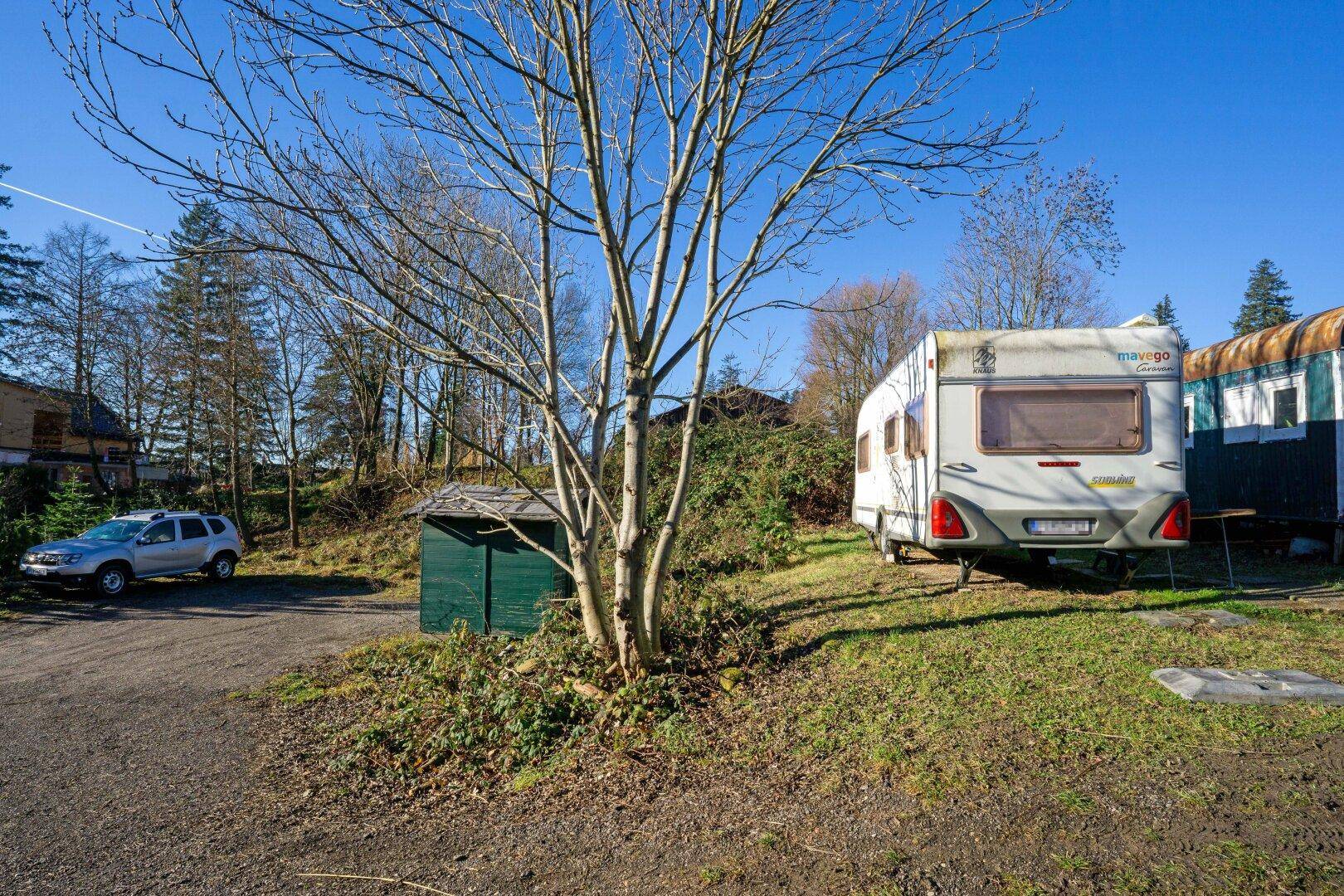 Außenbereich mit Kiesweg, einem Baum, einem kleinen Schuppen und einem geparkten Wohnwagen.