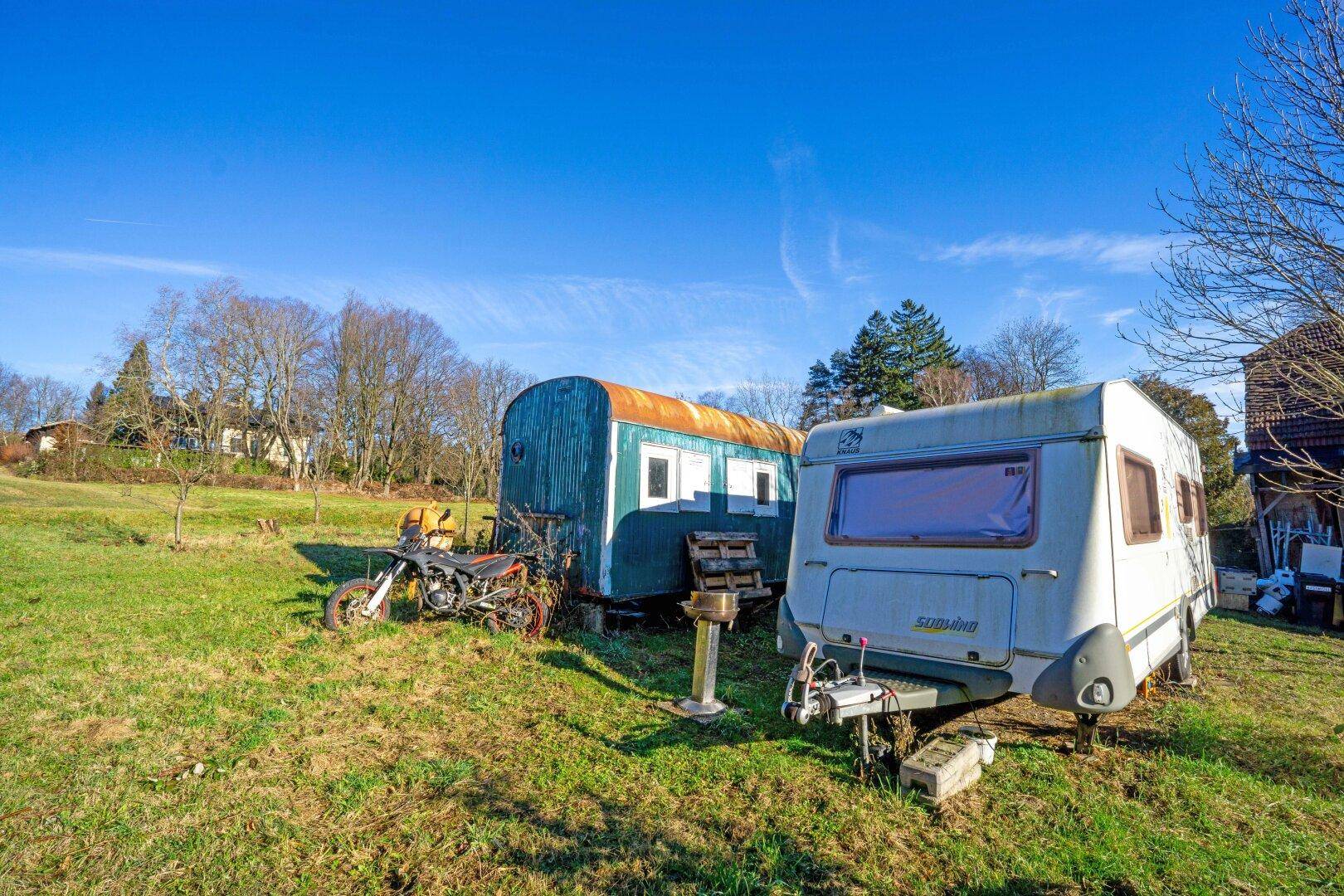 Grüne Wiese mit einem Wohnwagen, einem alten Bauwagen und einem Motorrad im Freien.