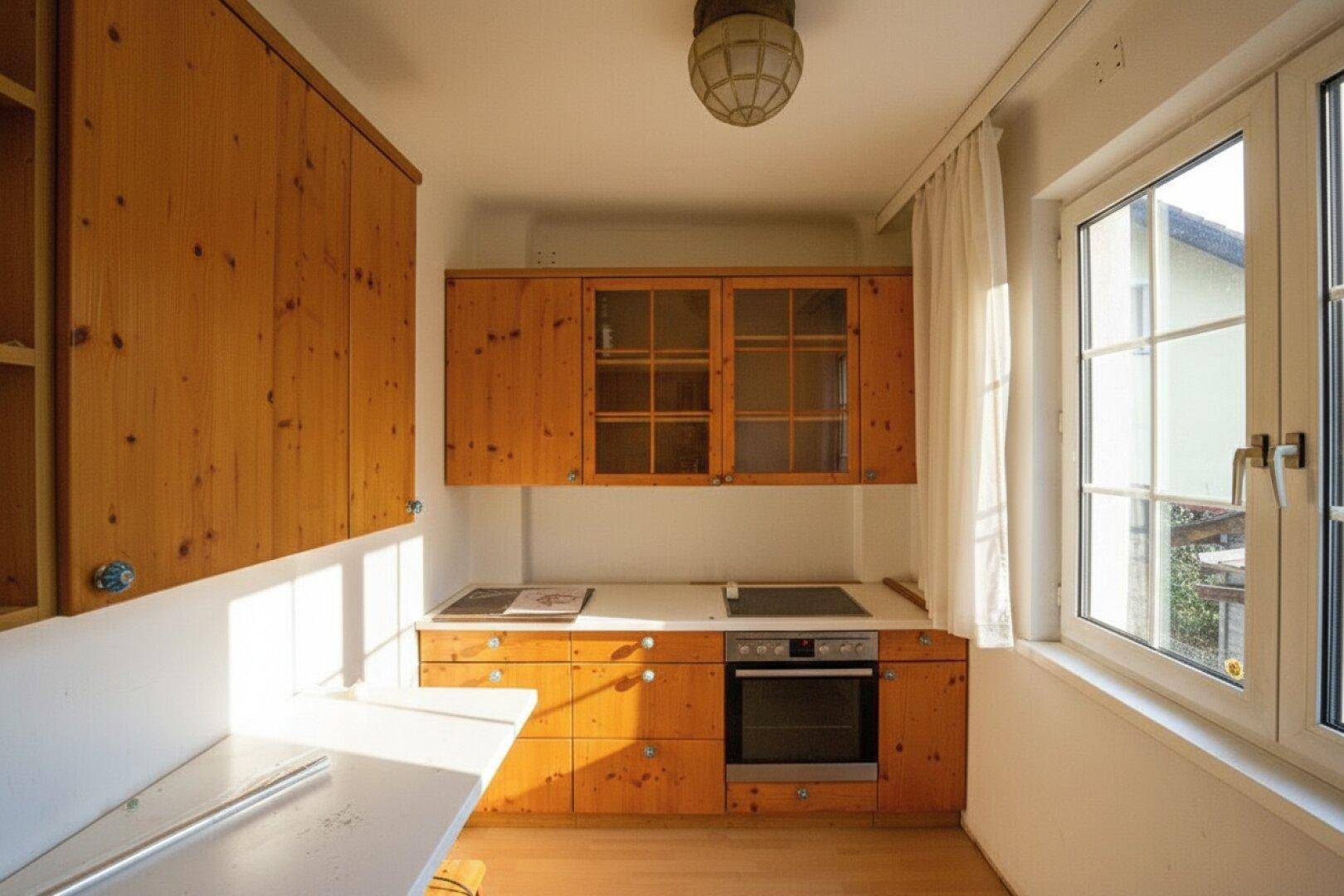 Kleine Küche mit Holzschränken, Einbauherd und Fenster, gut beleuchtet durch Sonnenlicht.