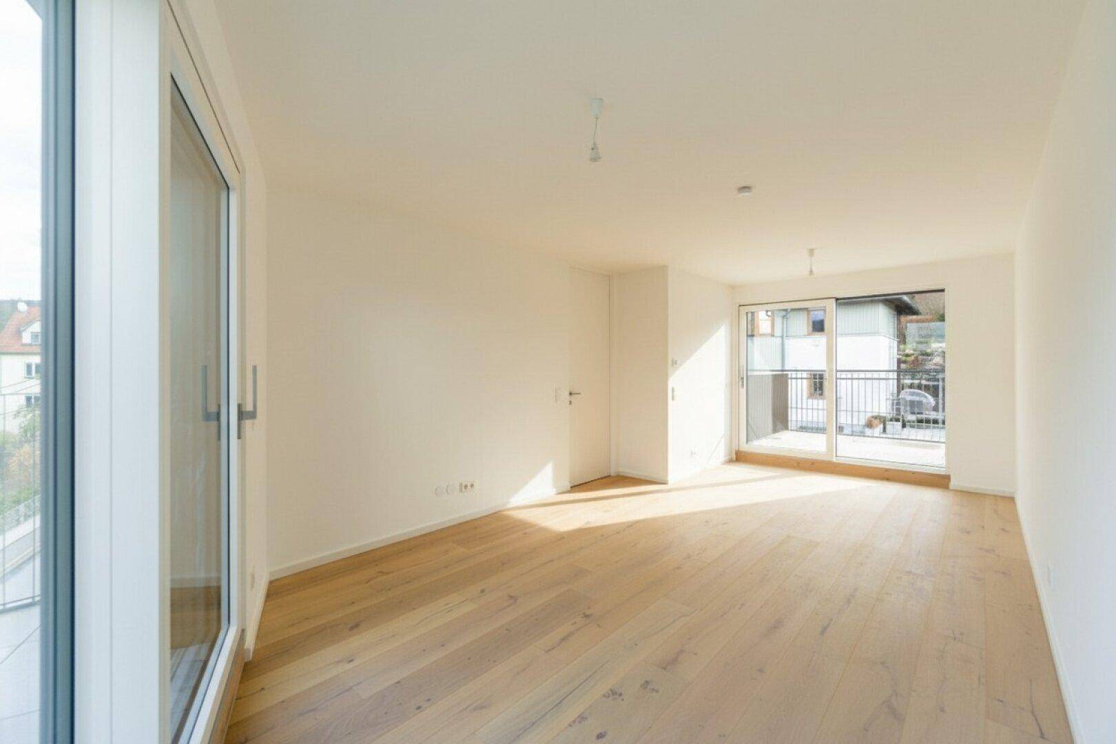 Sonniges Wohnzimmer mit hellem Holzboden, großen Fenstern und Zugang zu zwei Balkonen.