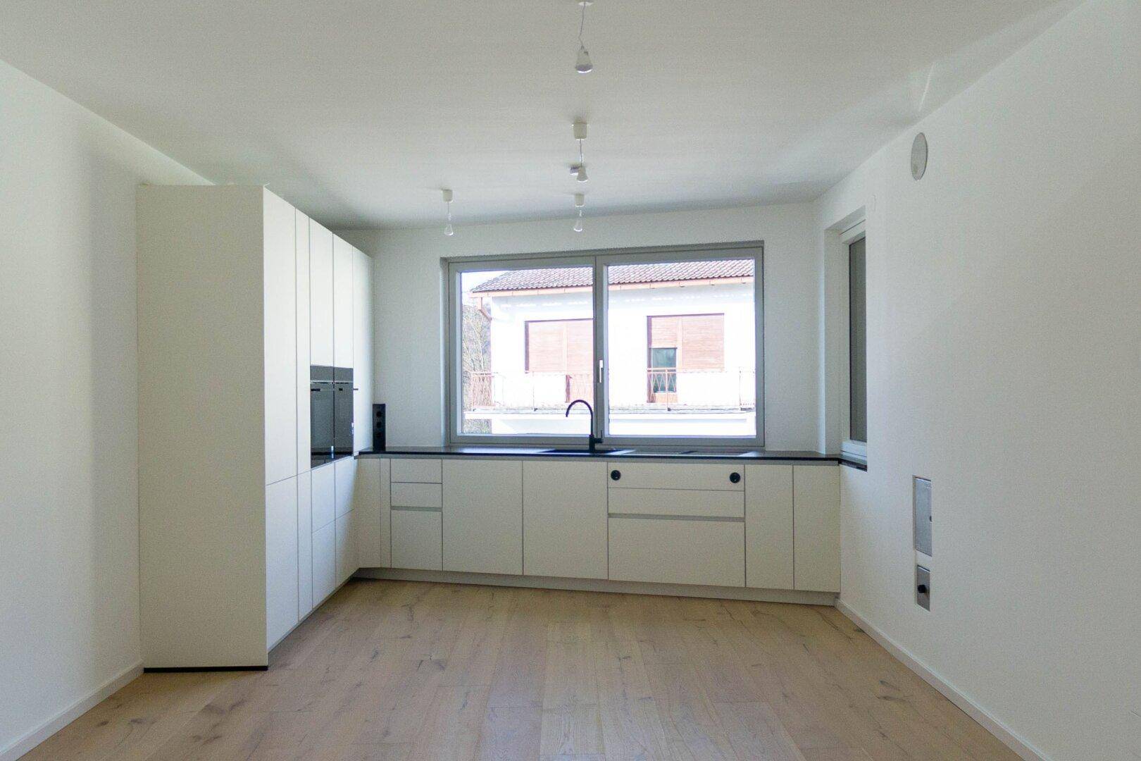 Moderne Einbauküche mit weißen Schränken, dunkler Arbeitsplatte und großem Fenster.