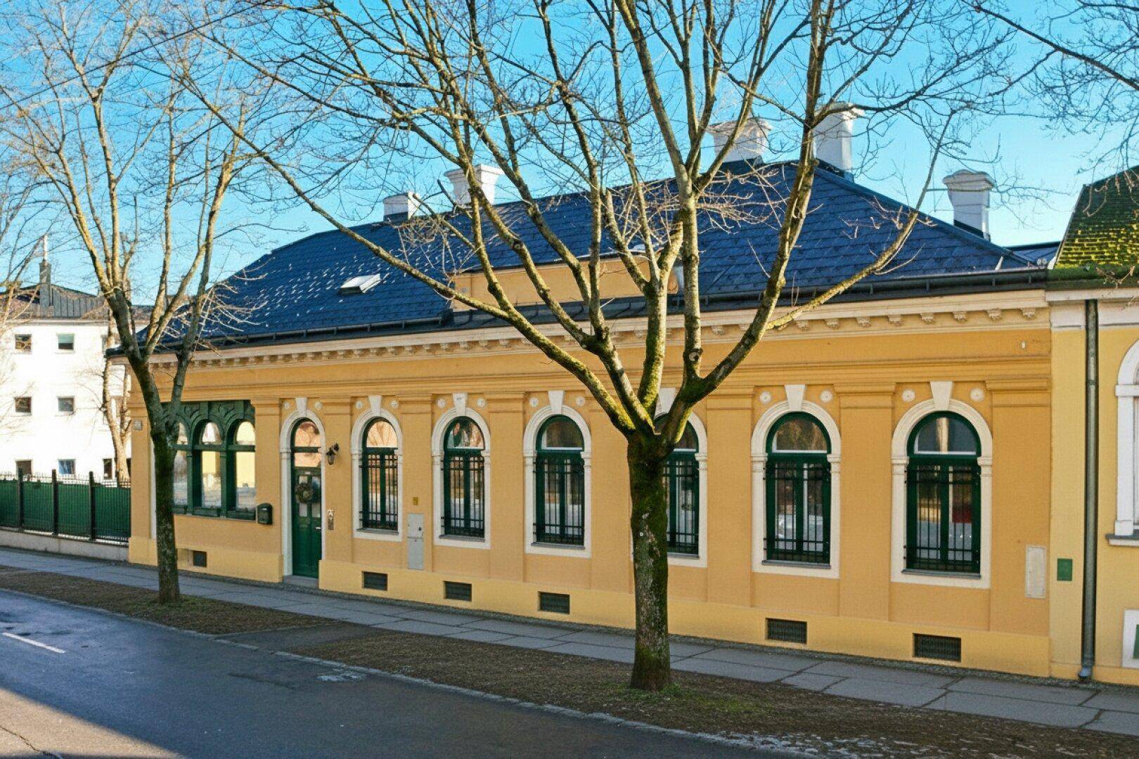 Historisches Gebäude mit gelber Fassade, grünen Fenstern und dunklem Dach, von der Straße aus gesehen.