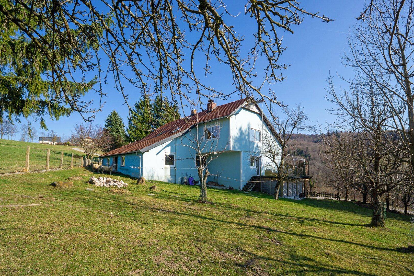 Blaues Haus mit rotem Dach, umgeben von Bäumen und grünen Wiesen in hügeliger Landschaft.