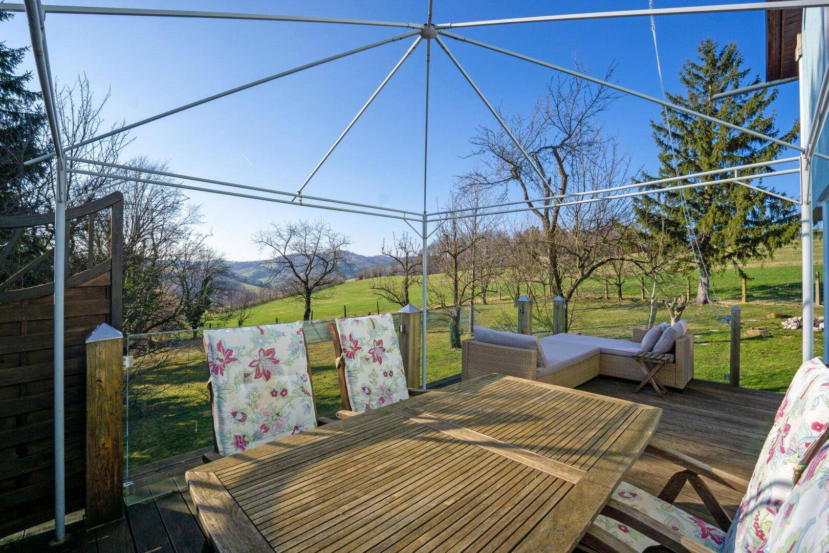 Überdachte Holzterrasse mit Esstisch und Liegestühlen, umgeben von grüner Natur.