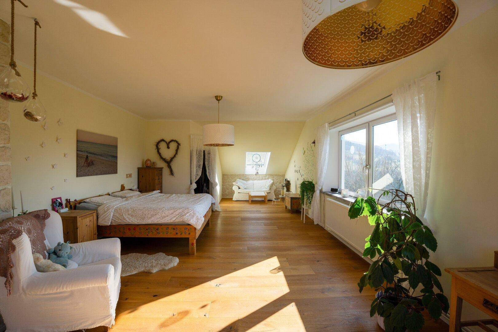 Helles Schlafzimmer mit Holzboden, großem Bett und gemütlicher Sitzecke unter dem Dachfenster.