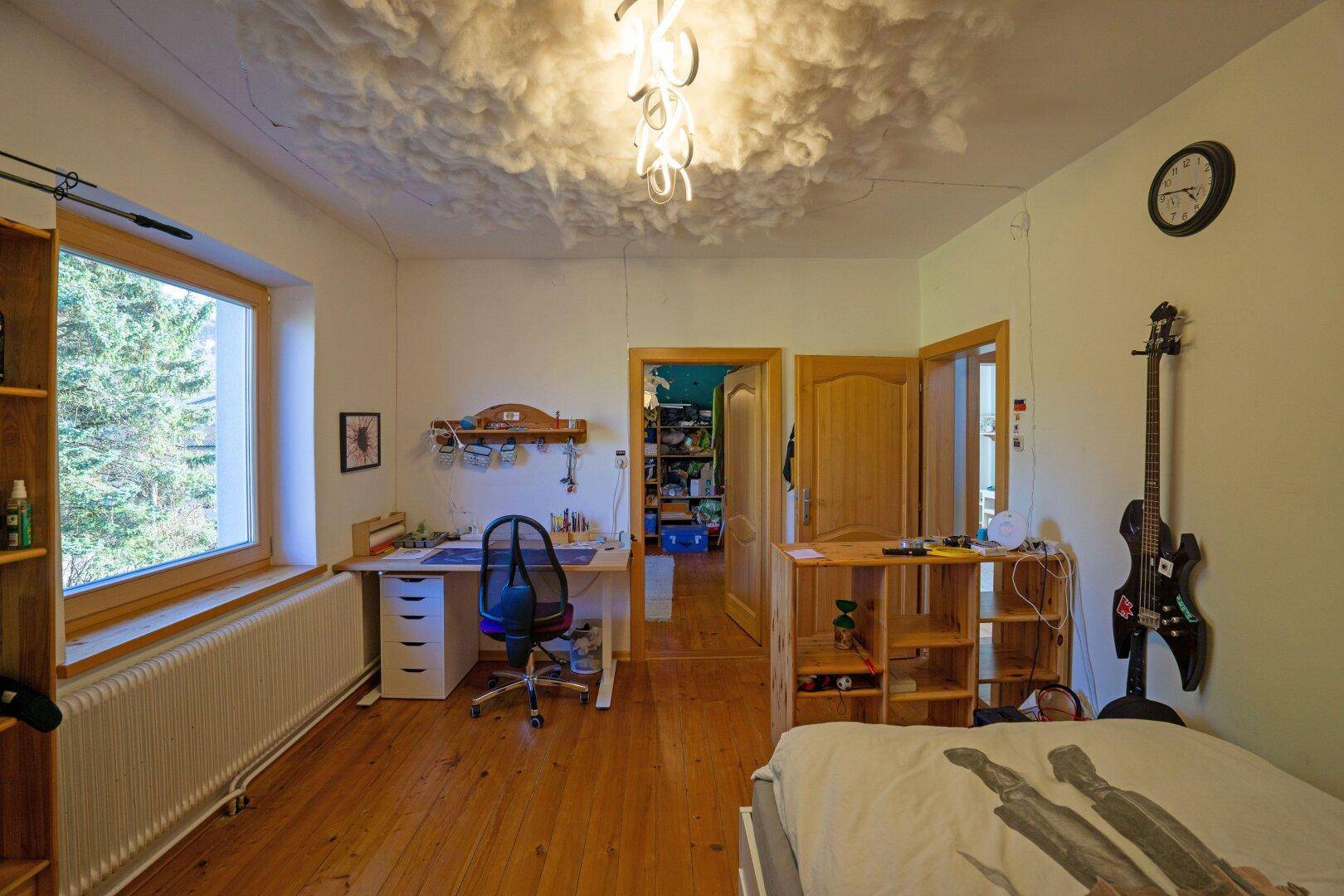 Schlafzimmer mit Holzboden, Schreibtisch und Fenster, das viel Tageslicht hereinlässt.