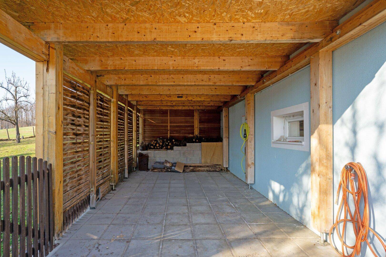 Überdachter Carport mit Holzkonstruktion und Lagerplatz für Brennholz.