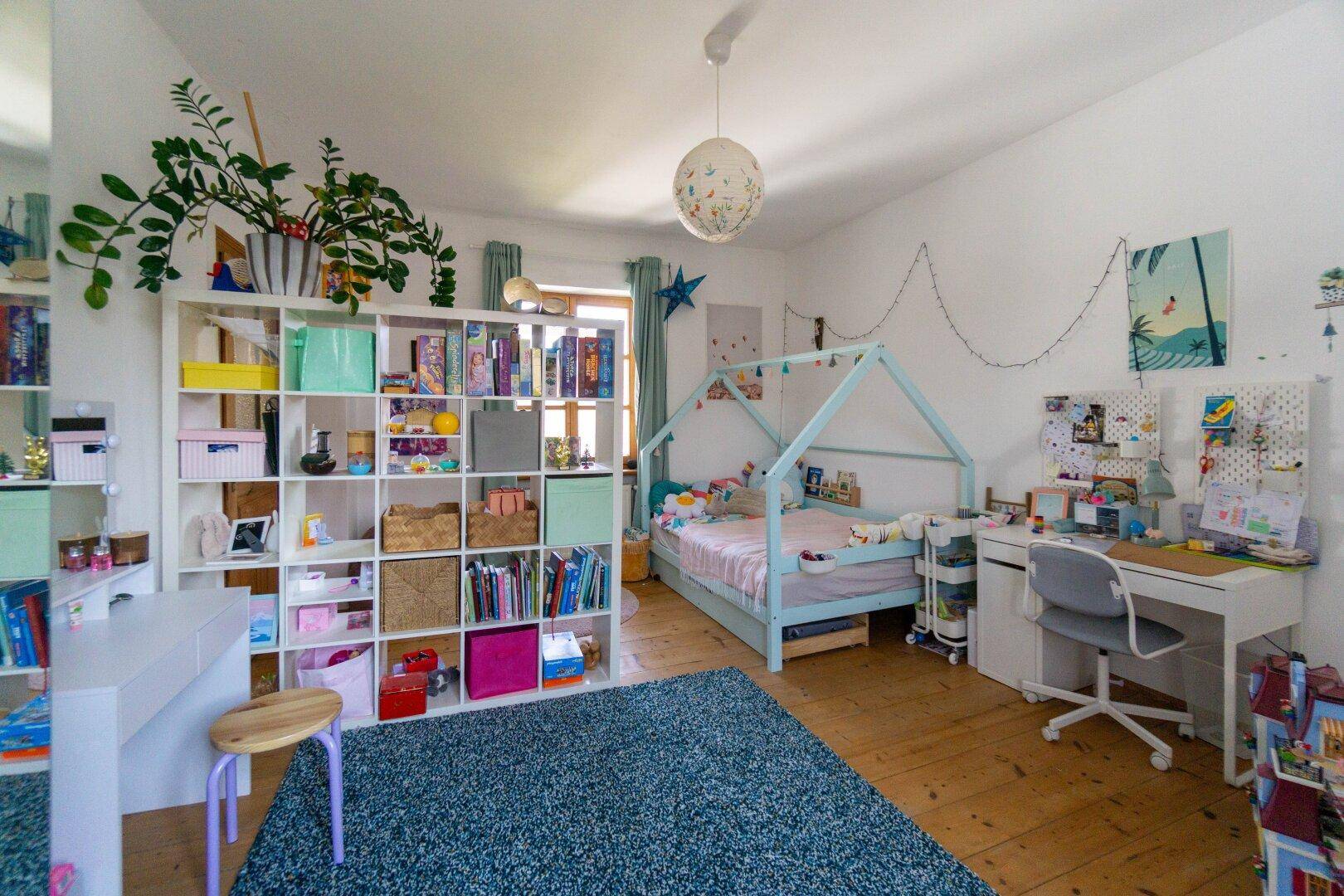 Helles Kinderzimmer mit Holzboden, großem Regal und farbenfroher Dekoration.