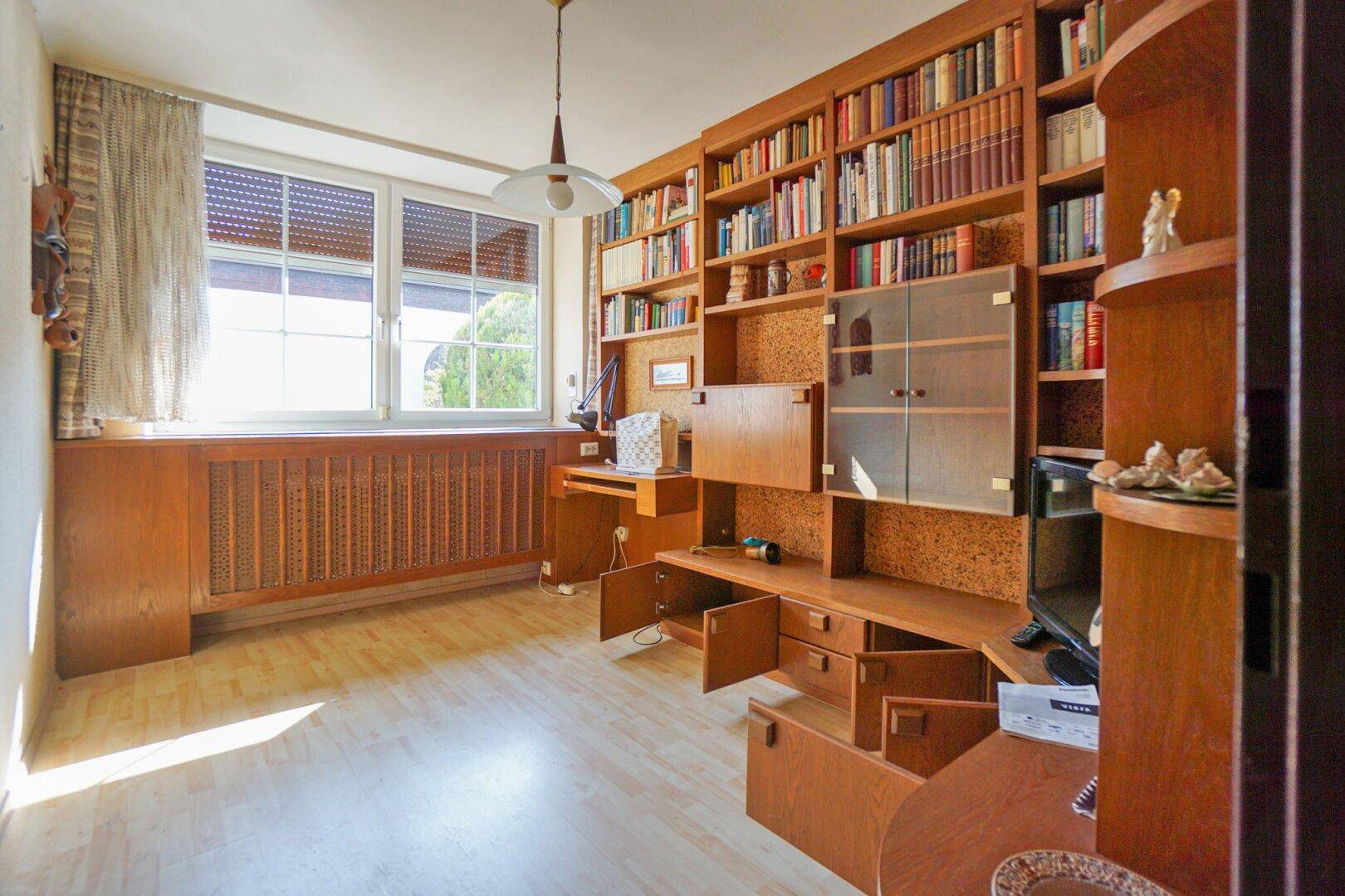Helles Arbeitszimmer mit großer Bücherwand aus Holz und einem Schreibtisch am Fenster.