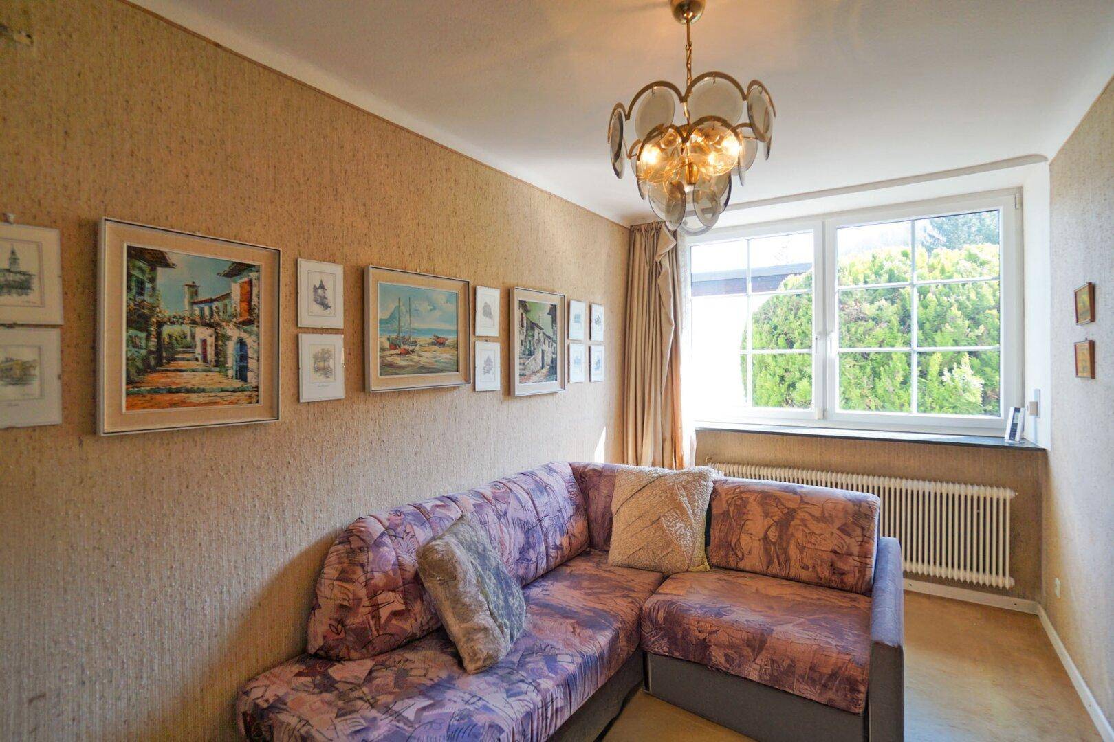 Gemütliches Wohnzimmer mit Ecksofa, Bildern an der Wand und einem Fenster mit Blick ins Grüne.
