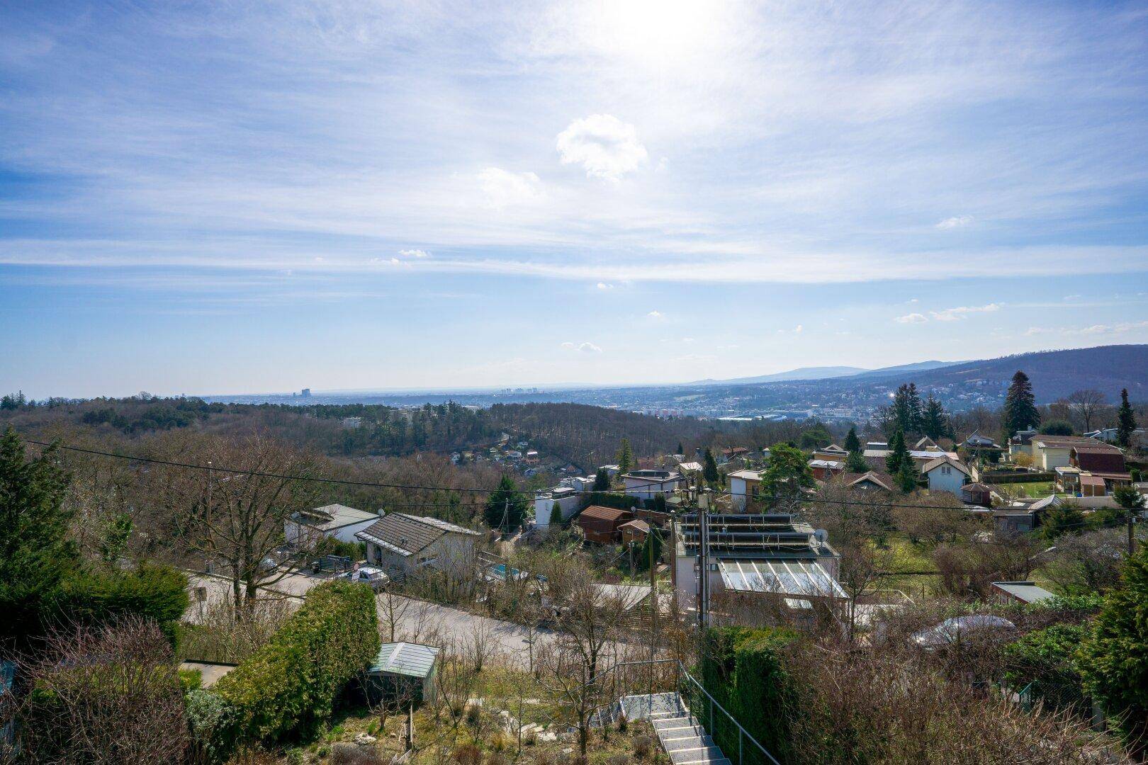 Weitläufiger Panoramablick über die grüne Landschaft mit Siedlungen und Bergen unter blauem Himmel.