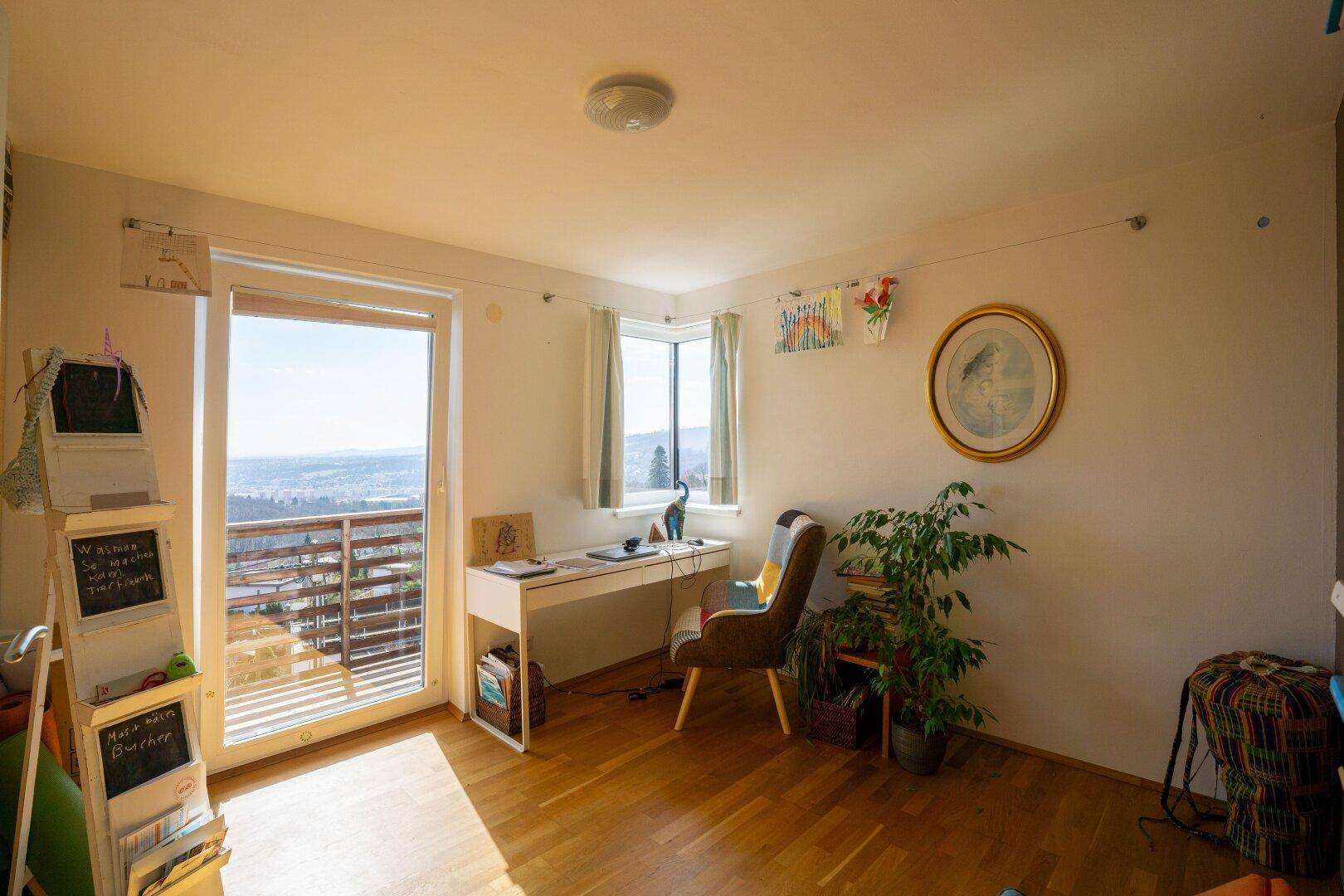 Helles Arbeitszimmer mit Schreibtisch, Pflanzen und großem Fenster mit weitem Blick ins Grüne.