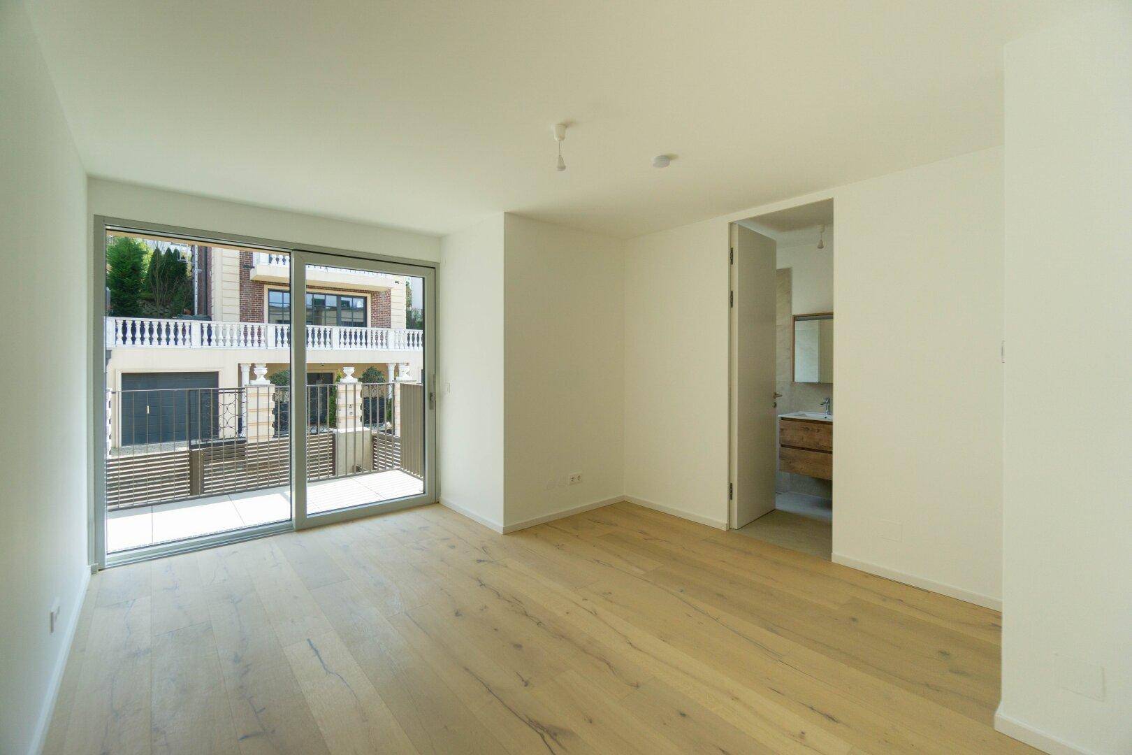 Helles Zimmer mit Holzboden und Balkonzugang, angrenzend an ein modernes Badezimmer.