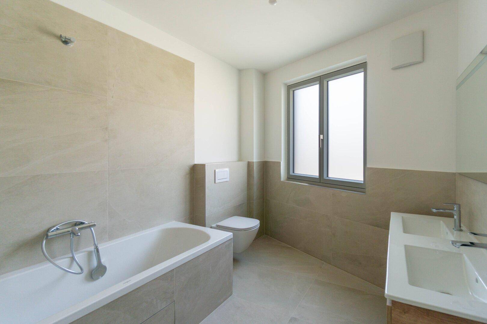 Modernes Badezimmer mit beigefarbenen Fliesen, Badewanne, wandhängendem WC und Doppelwaschbecken.