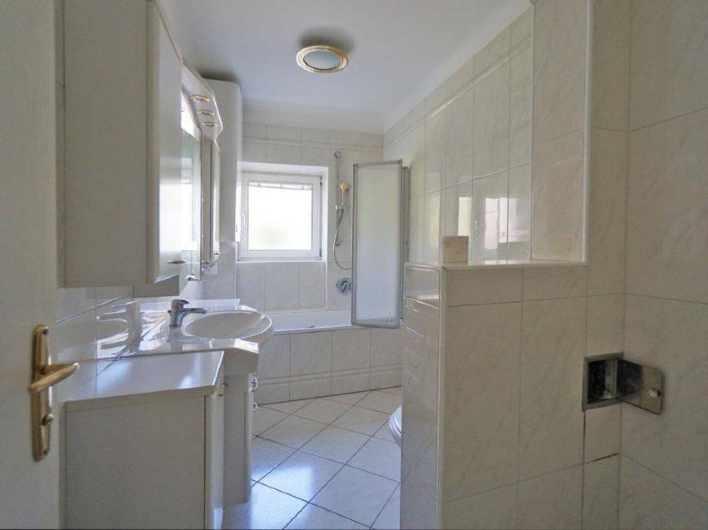 Gefliestes Badezimmer mit Badewanne, Duschabtrennung, Fenster und Waschtisch mit Spiegelschrank.