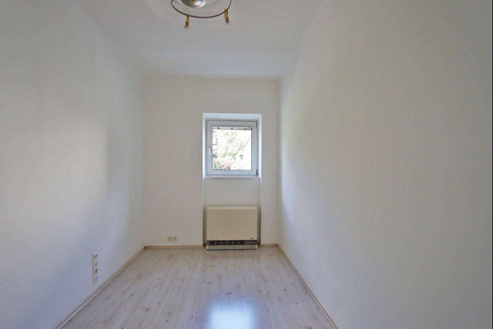 Leeres, helles Zimmer mit Laminatboden und einem Fenster mit Blick ins Grüne.