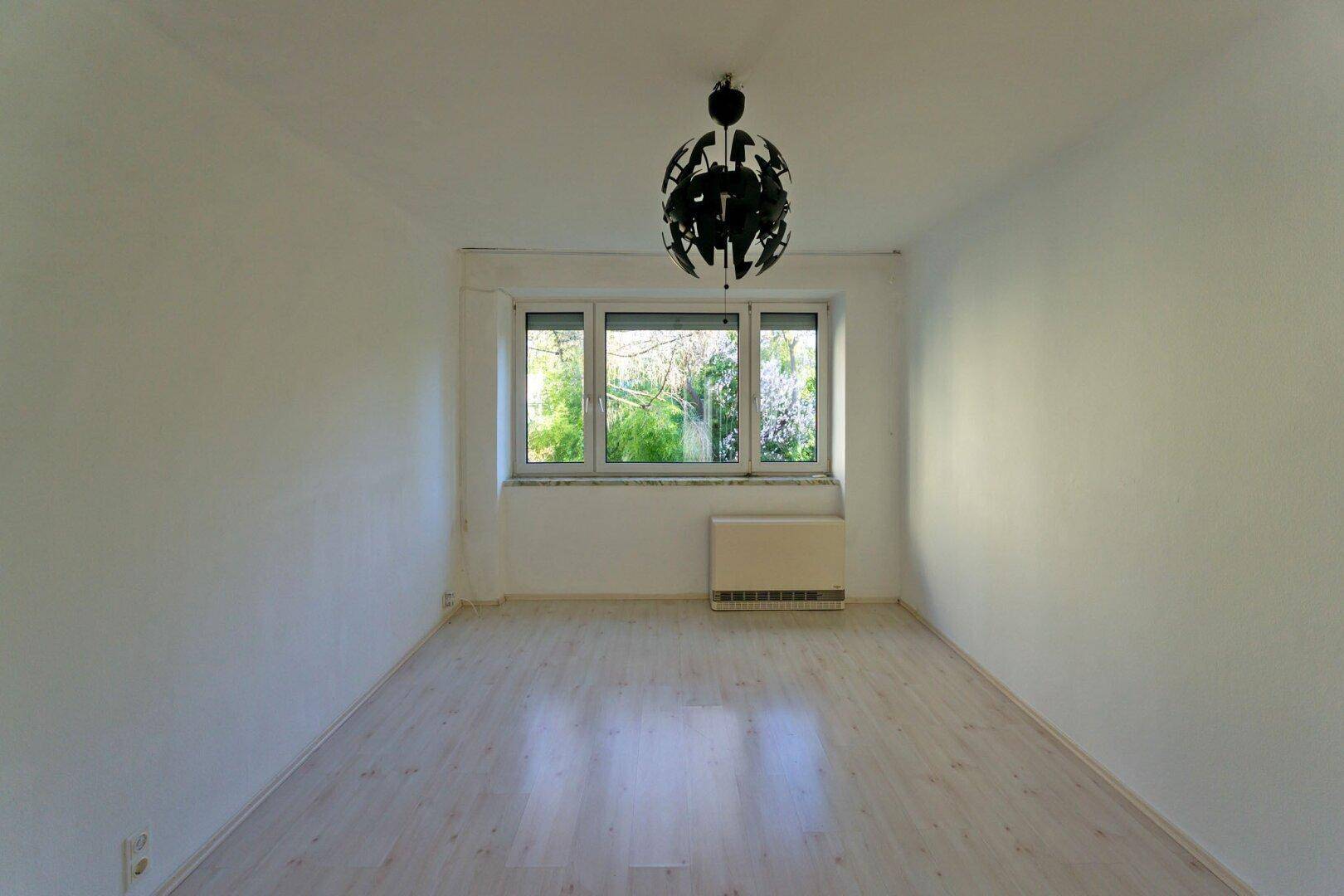 Heller, leerer Raum mit Laminatboden und großem Fenster mit Blick auf Bäume.