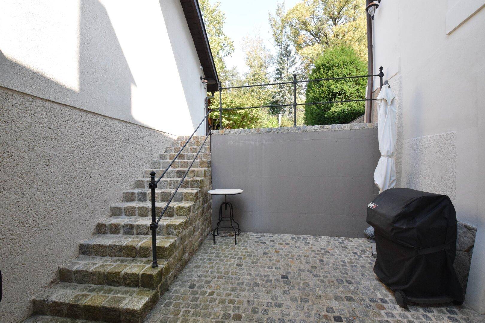 Steintreppe führt zu einem gepflasterten Bereich mit Grill, umgeben von Natur.