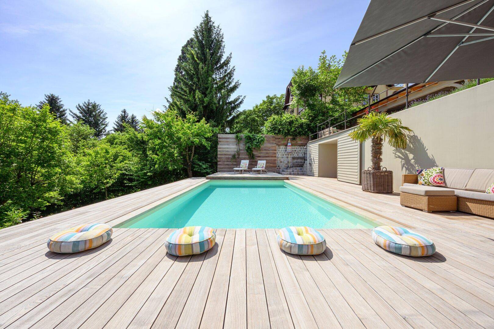 Moderner Poolbereich mit Holzdeck und farbenfrohen Kissen, umgeben von grüner Natur.