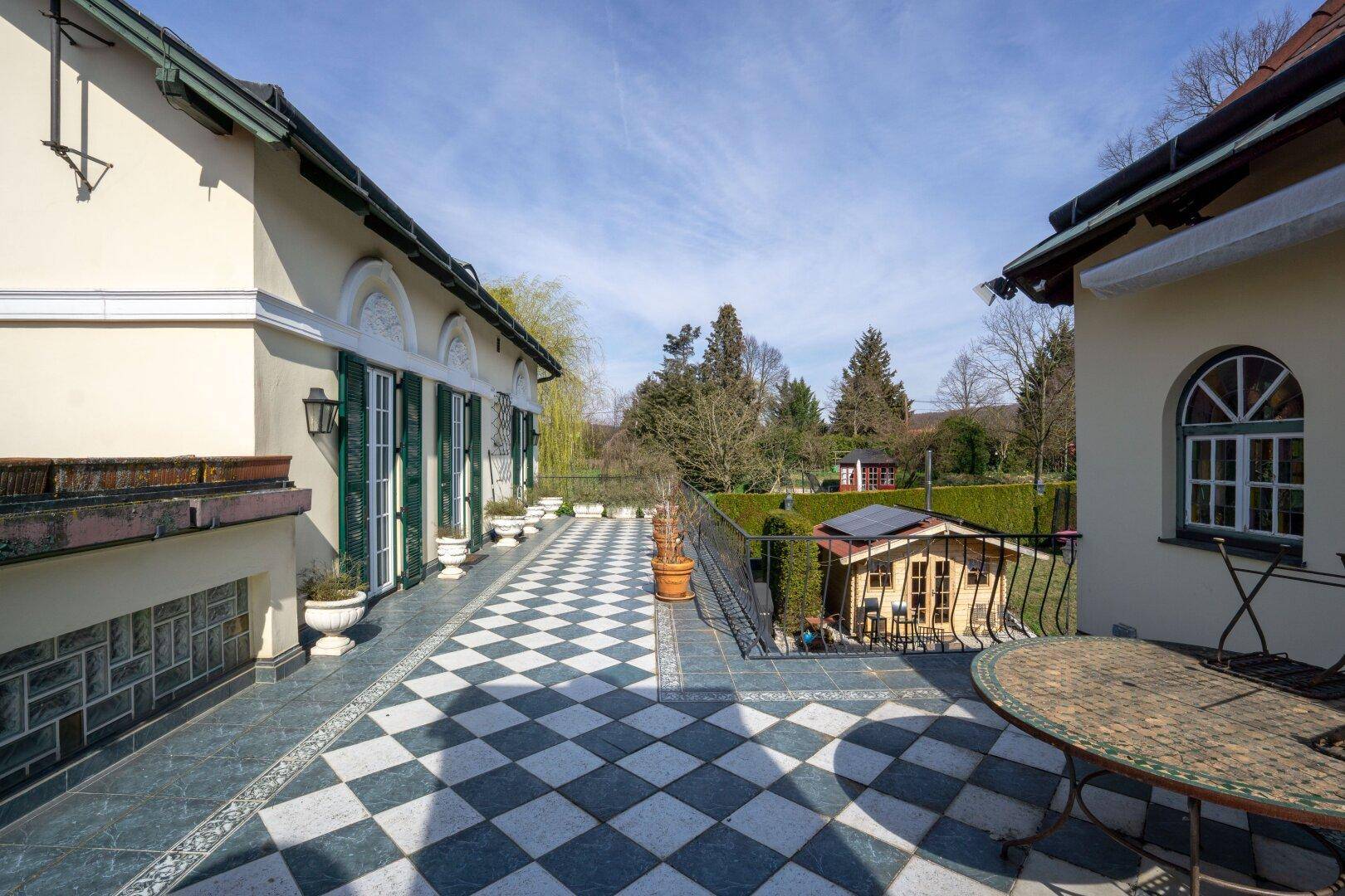 Geräumige Terrasse mit schwarz-weißem Fliesenmuster und Blick auf das Gartenhaus und die grüne Umgebung.