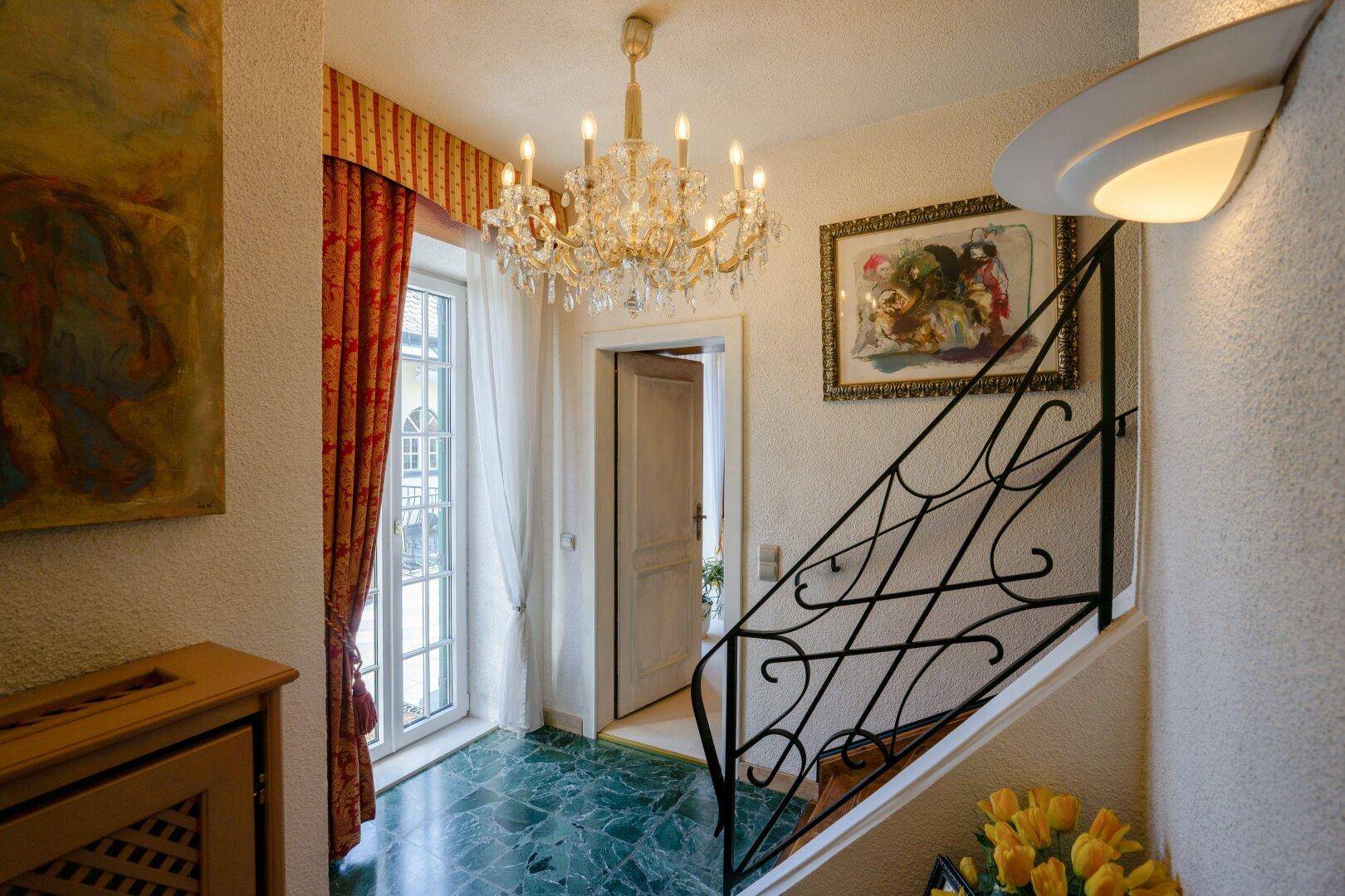 Heller Flur mit Marmorboden, Kronleuchter und Zugang zu einem Treppenhaus mit schmiedeeisernem Geländer.