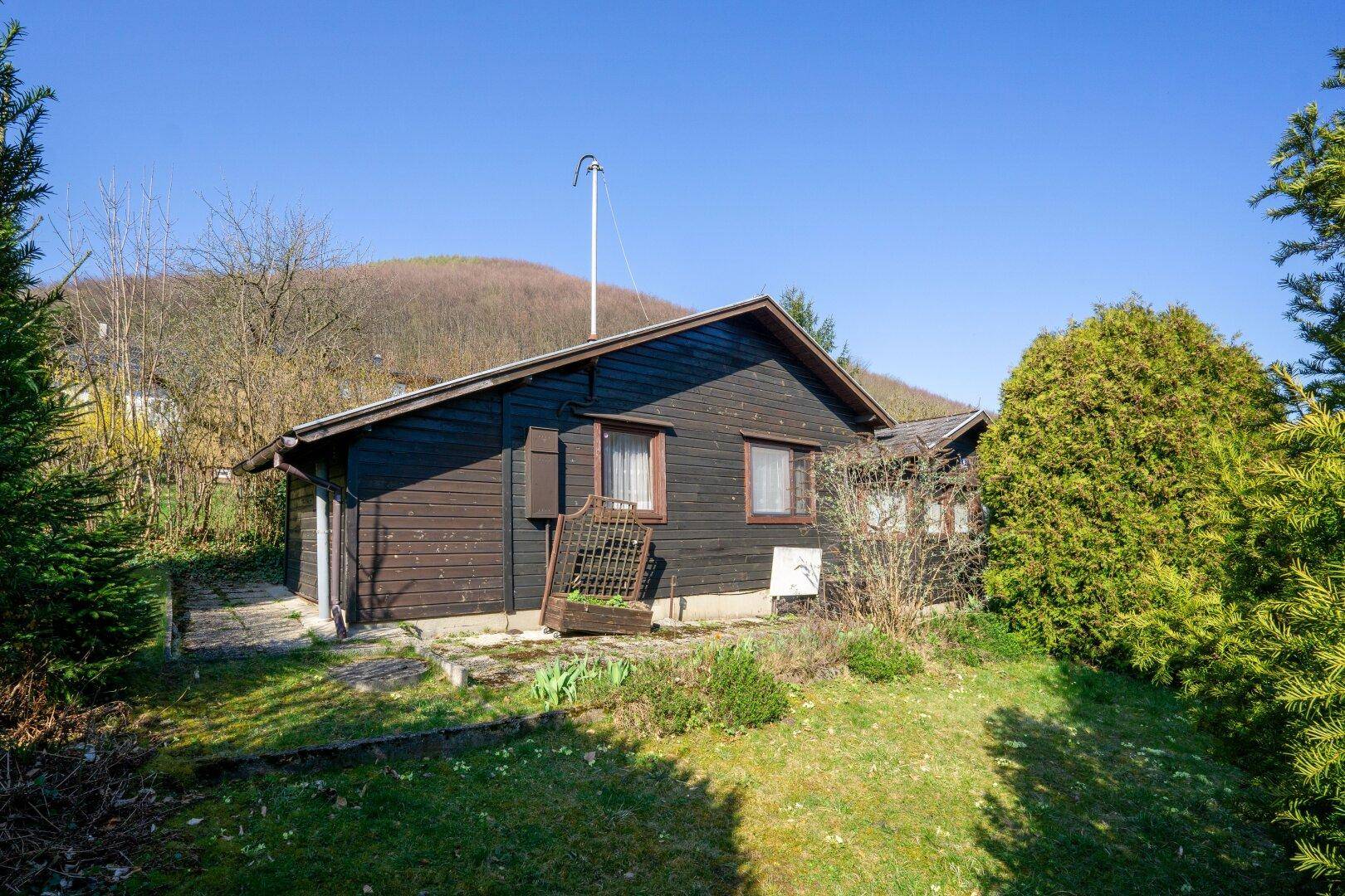 Ansicht des Holzhauses vom Garten aus, umgeben von Rasen und Sträuchern unter blauem Himmel.