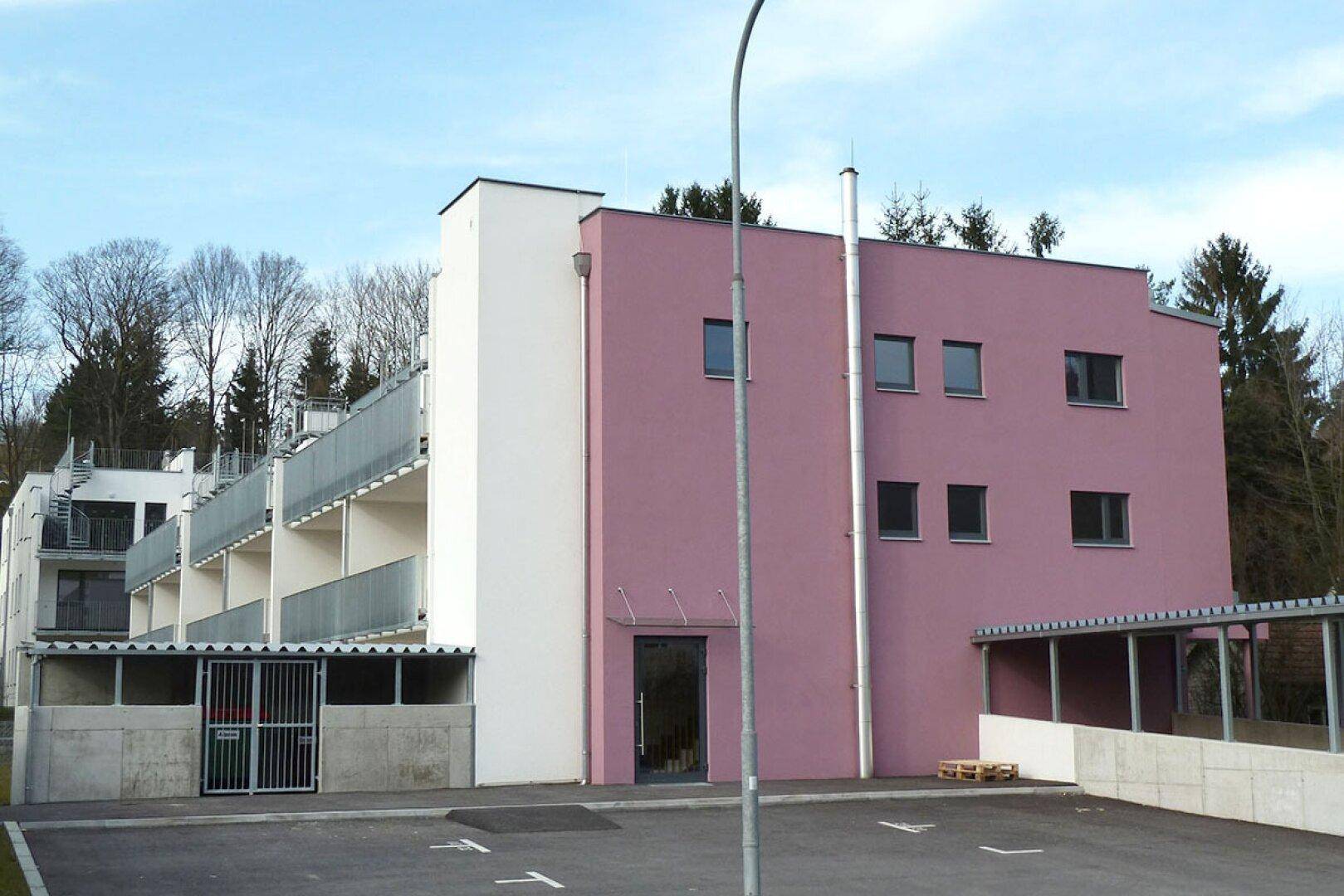 Moderne Gebäudefassade in Rosa und Weiß mit mehreren Balkonen und umgebenden Bäumen.