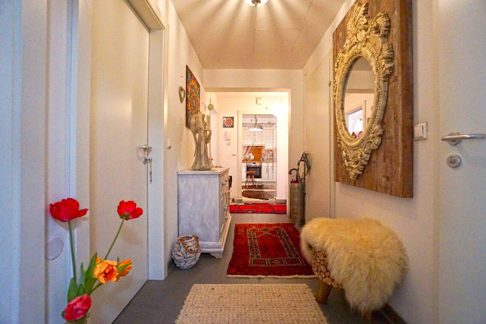 Heller Flur mit dekorativem Spiegel, Teppichen und Blumen, der zu weiteren Räumen führt.