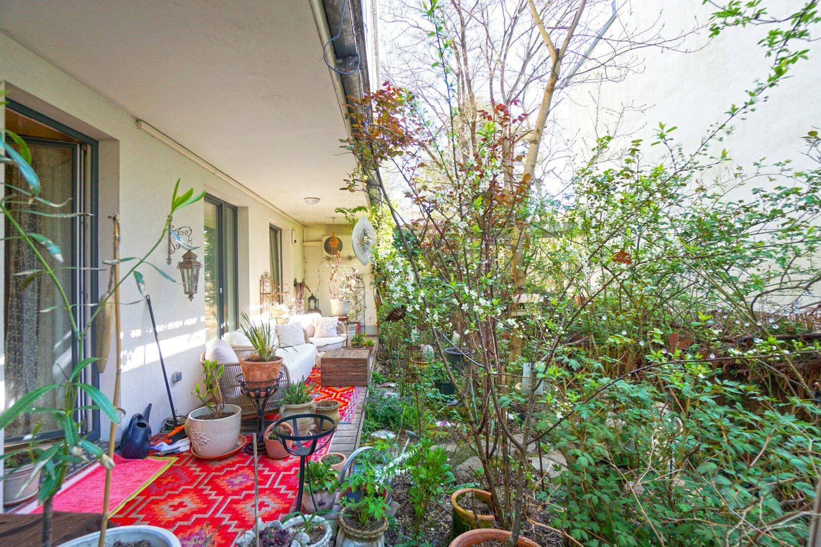 Lange Terrasse mit Sitzgelegenheiten und vielen Pflanzen, die in einen grünen Garten übergeht.